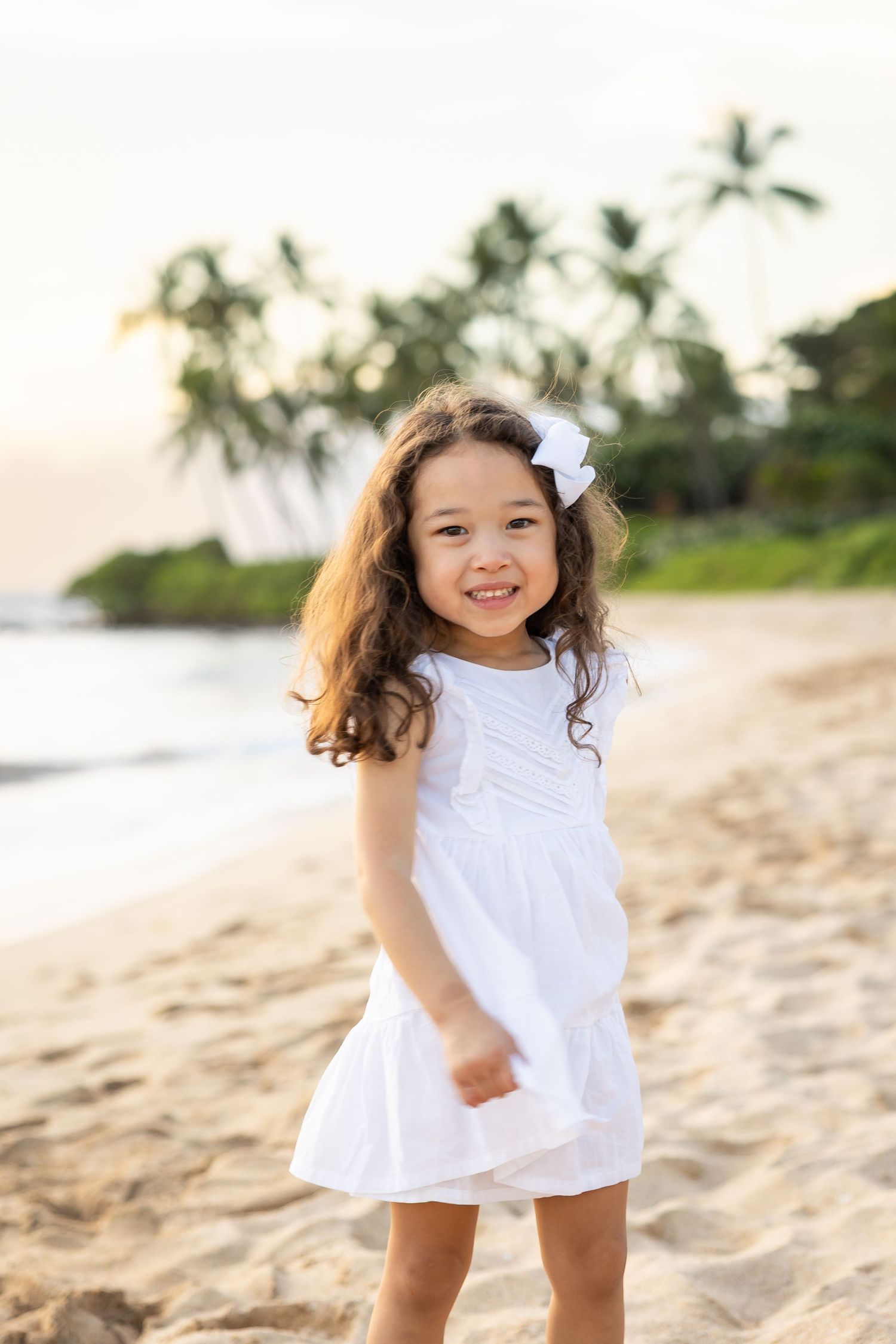 Young girl in white dress with bow smiles on sandy beach with palm trees at sunset.