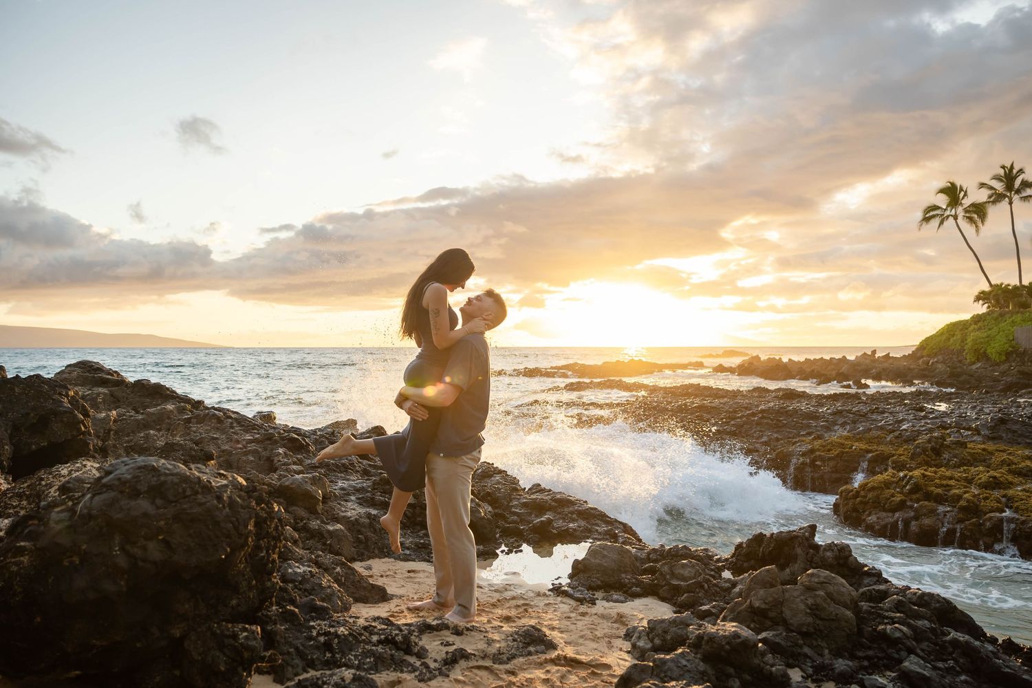 Couple embracing on rocky Hawaiian beach with dramatic crashing waves at sunset.