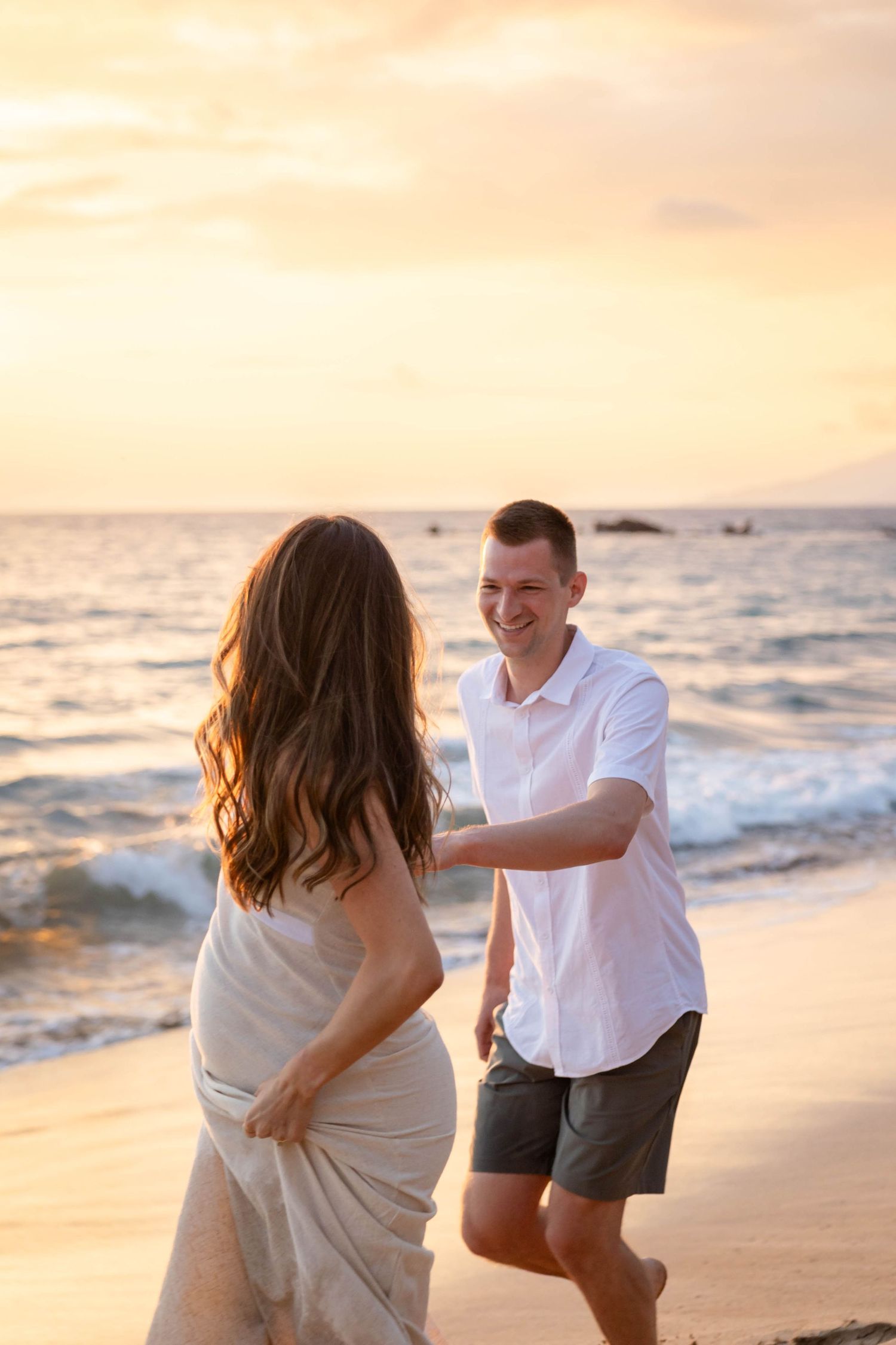 Smiling man in white shirt reaching for pregnant partner on beach during vibrant golden sunset.
