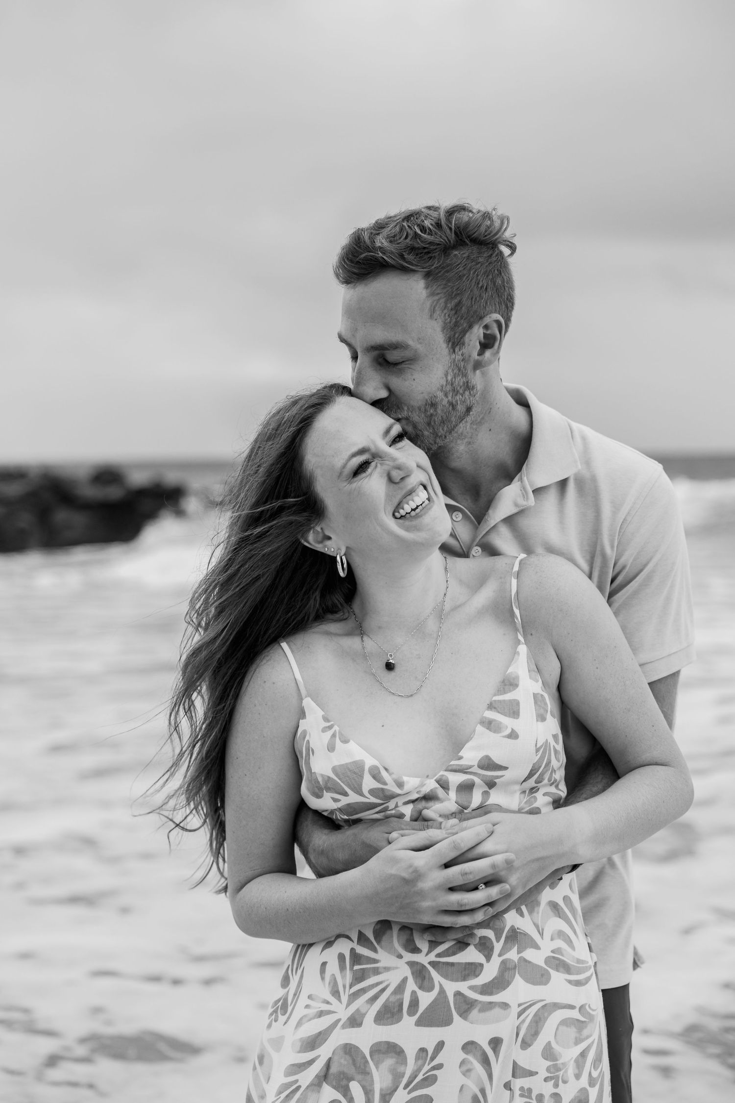 Black and white photo of laughing woman being embraced and kissed by man on rocky beach.