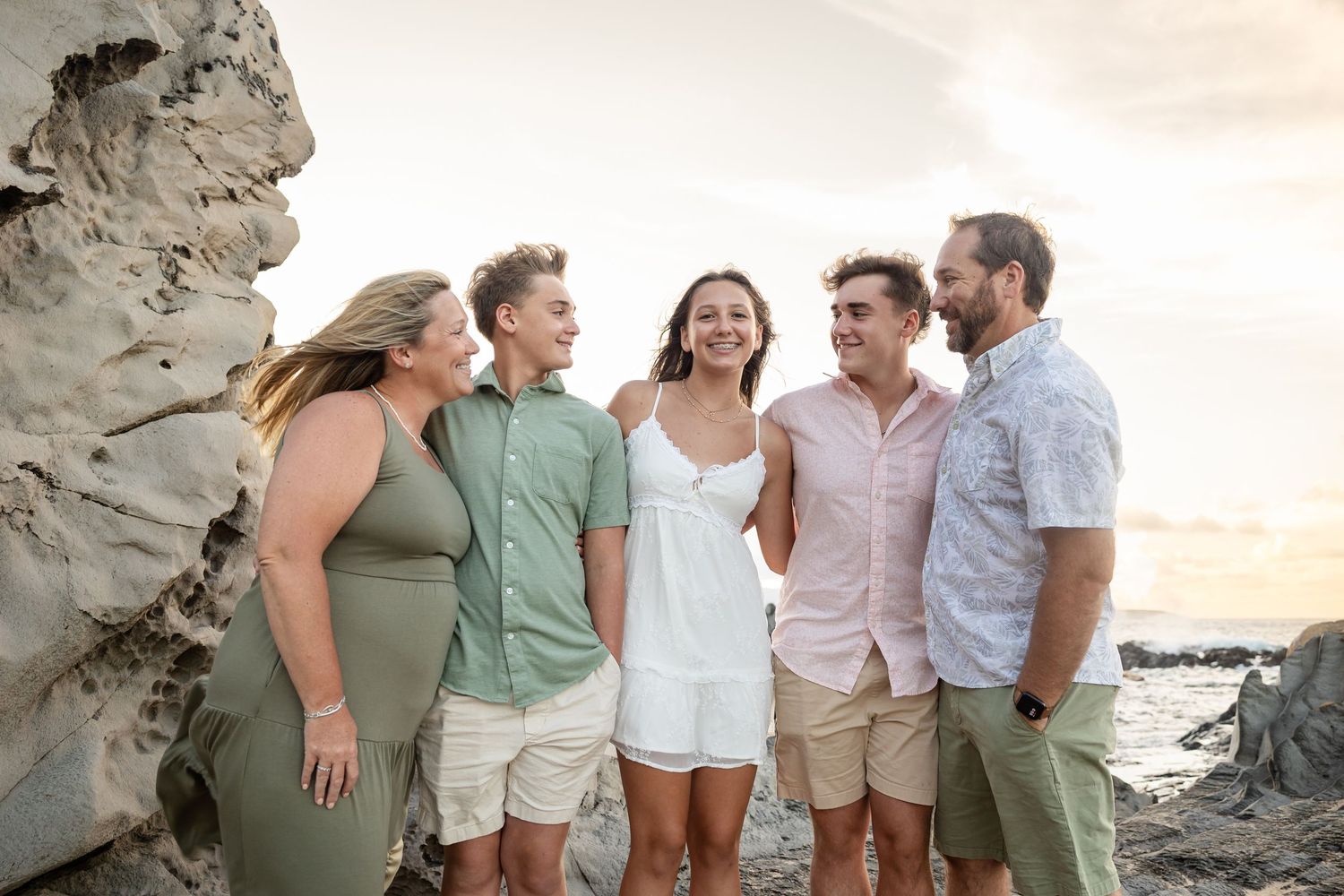 Family of five laughing together on a rocky Hawaii beach at sunset.