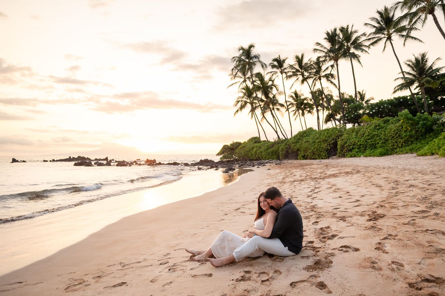 Couple sitting closely together on sandy tropical beach at sunset with palm trees behind them.