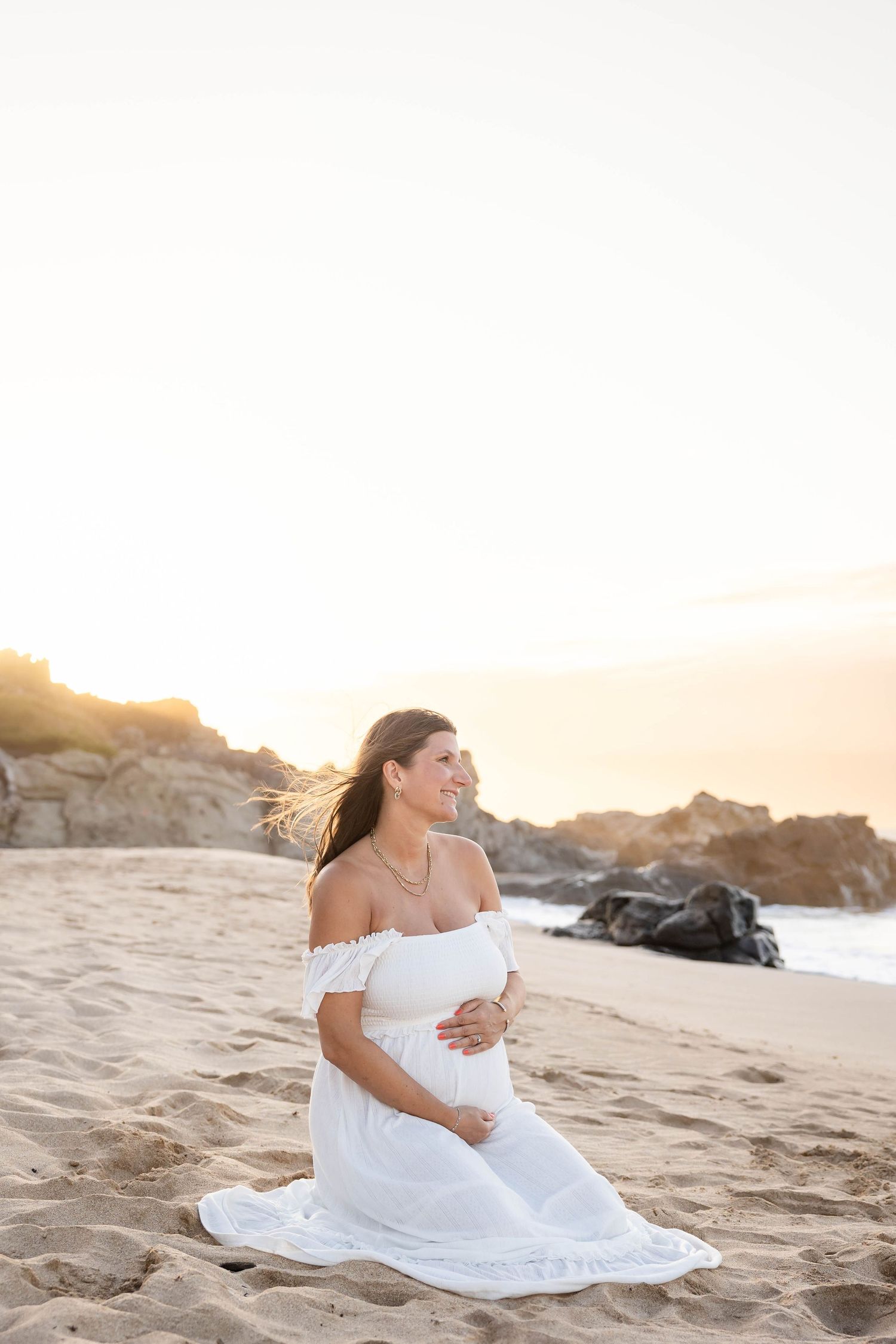 Glowing pregnant woman in white dress sitting on sandy beach at sunset.