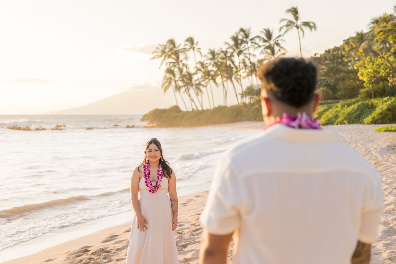 Couple wearing leis on a Hawaiian beach at sunset with palm trees and mountains in the background.