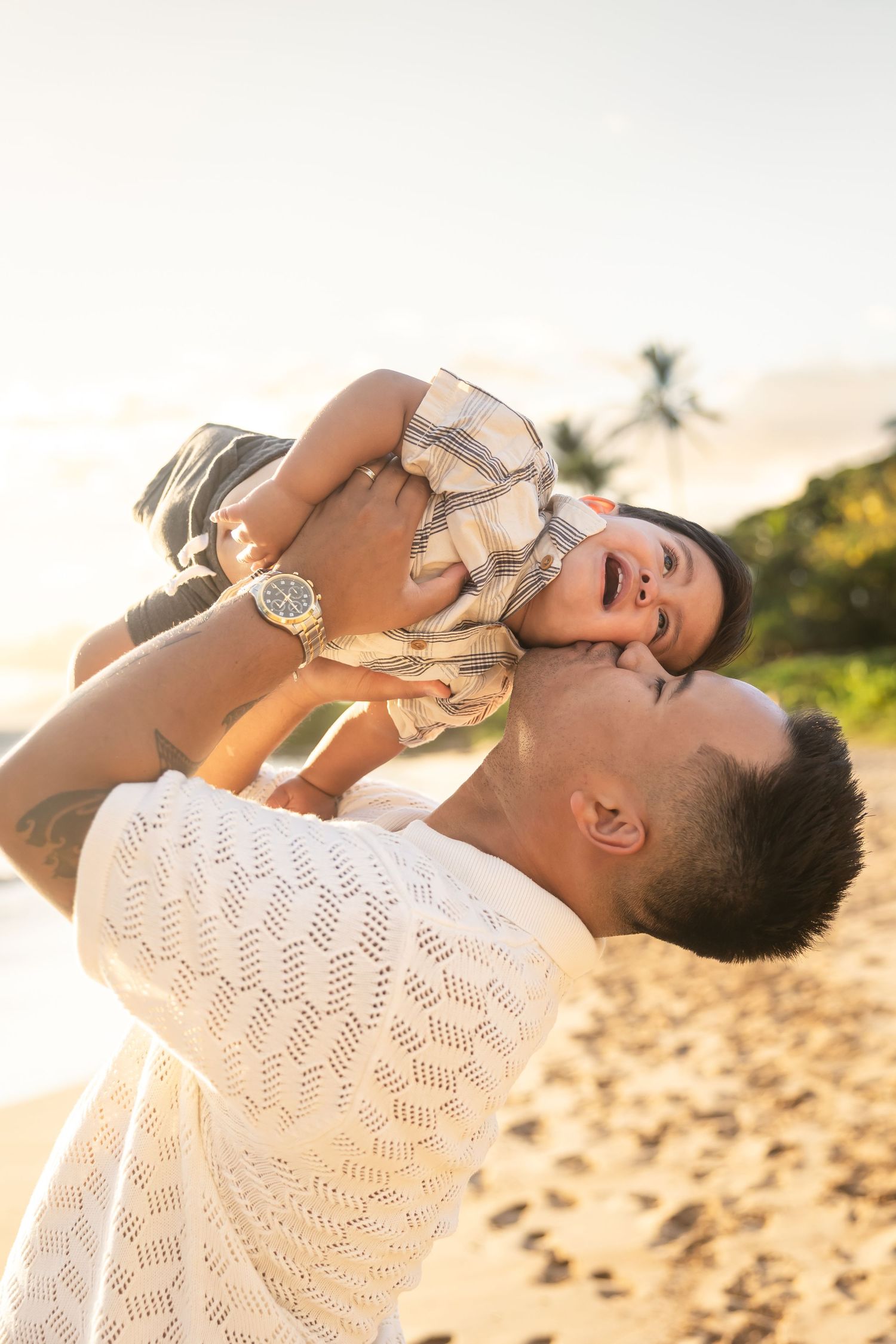 Father lifting his laughing baby boy overhead on a golden sunset beach with palm trees.