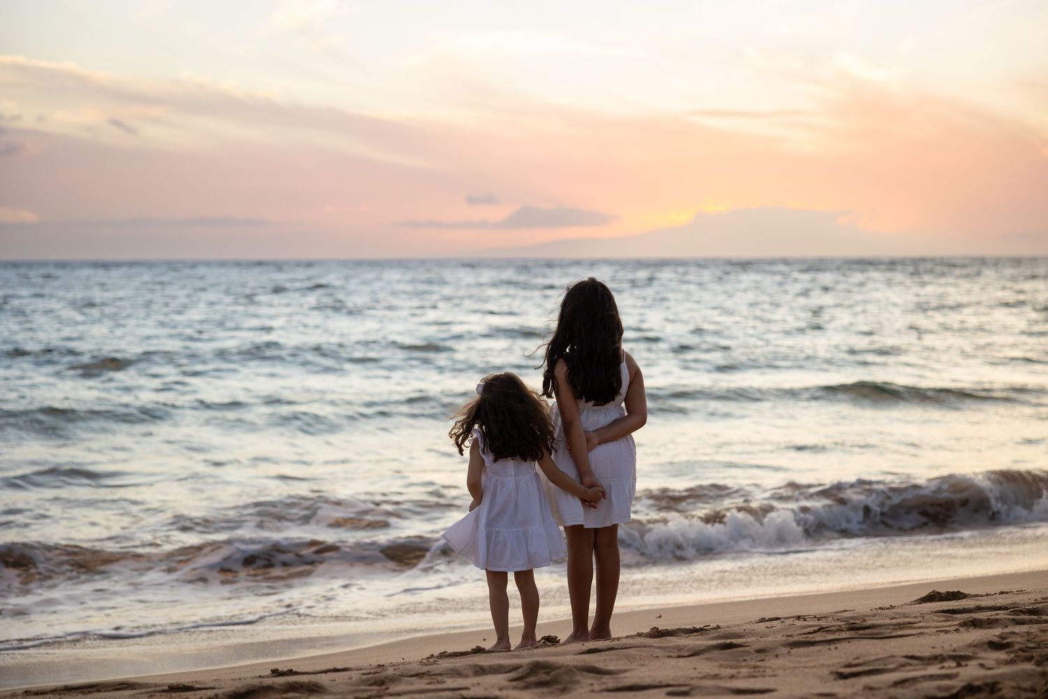 Two girls in white dresses hold hands on a sandy beach at sunset, gazing at the ocean waves.