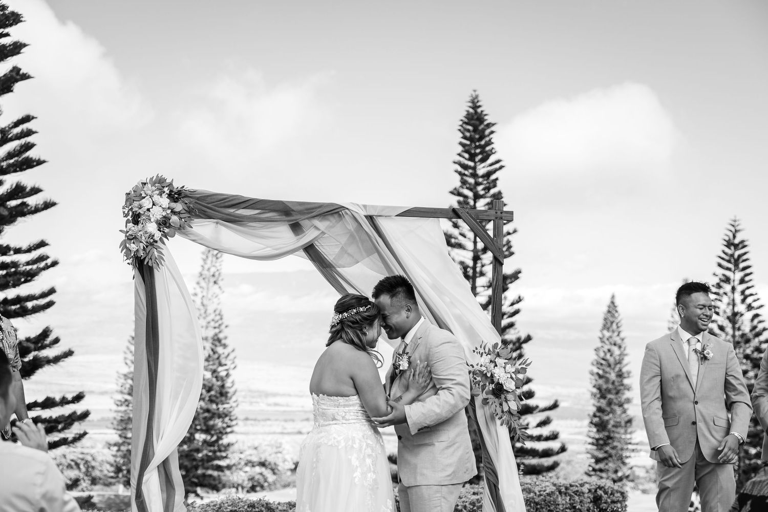 Black and white outdoor wedding ceremony under floral arch with pine trees and guests seated.