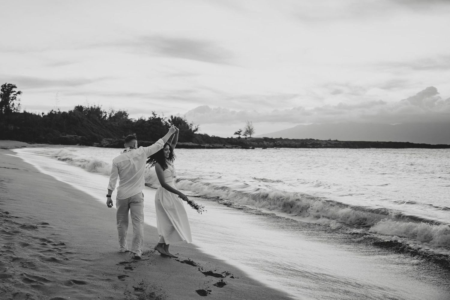 Black and white photo of a couple walking along a beach shoreline with waves and palm trees.