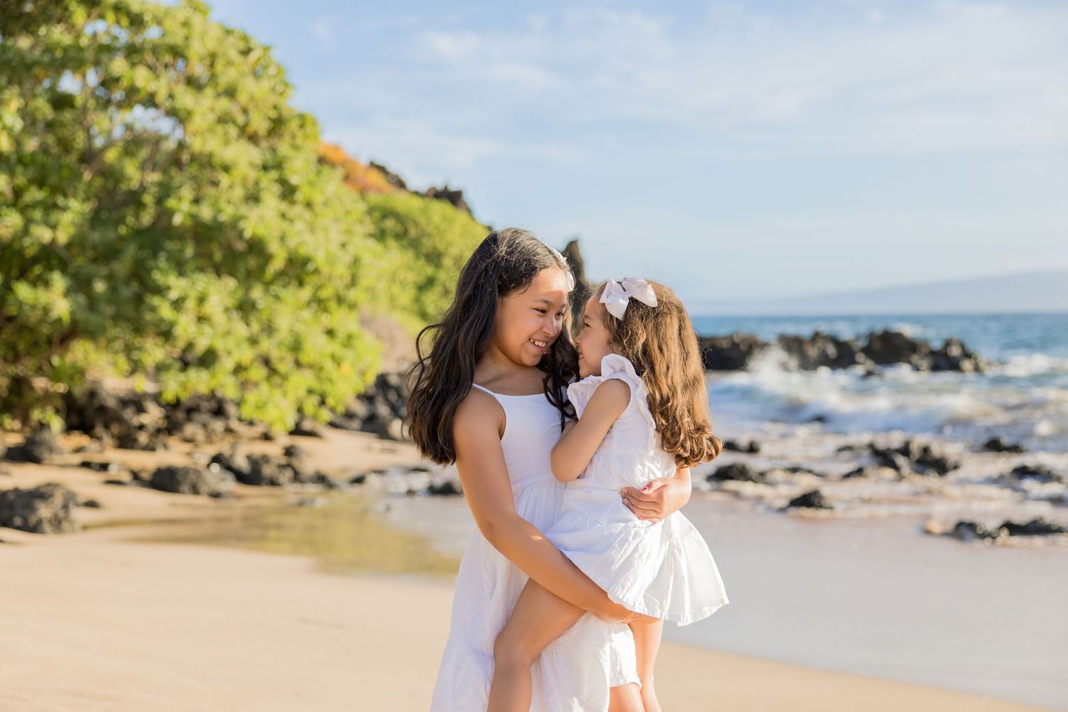 A mother and young daughter in white dresses share a tender embrace on a sunny Hawaiian beach.
