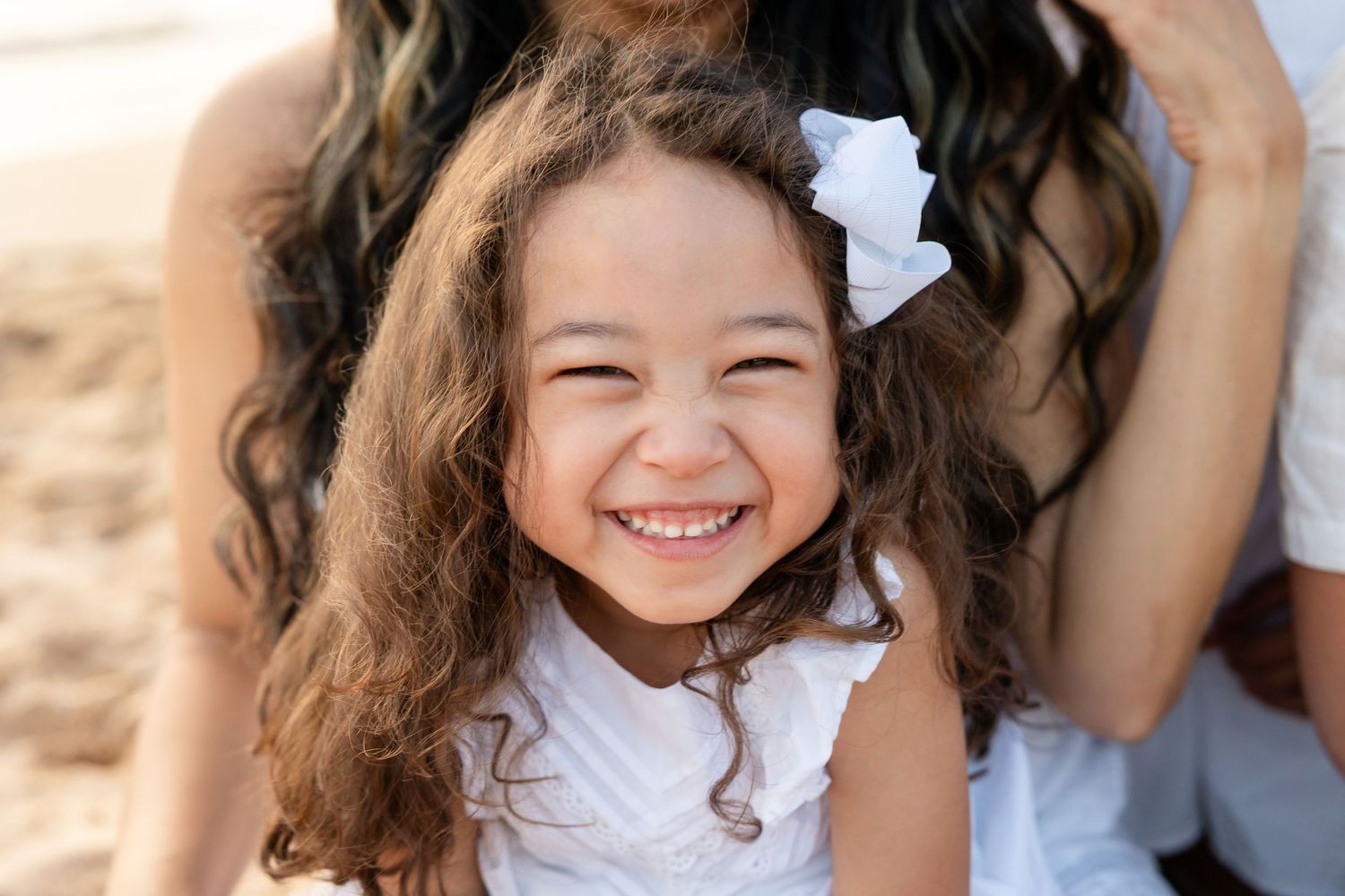 A joyful young girl with curly hair and a white bow smiles brightly, wearing a white dress outdoors.
