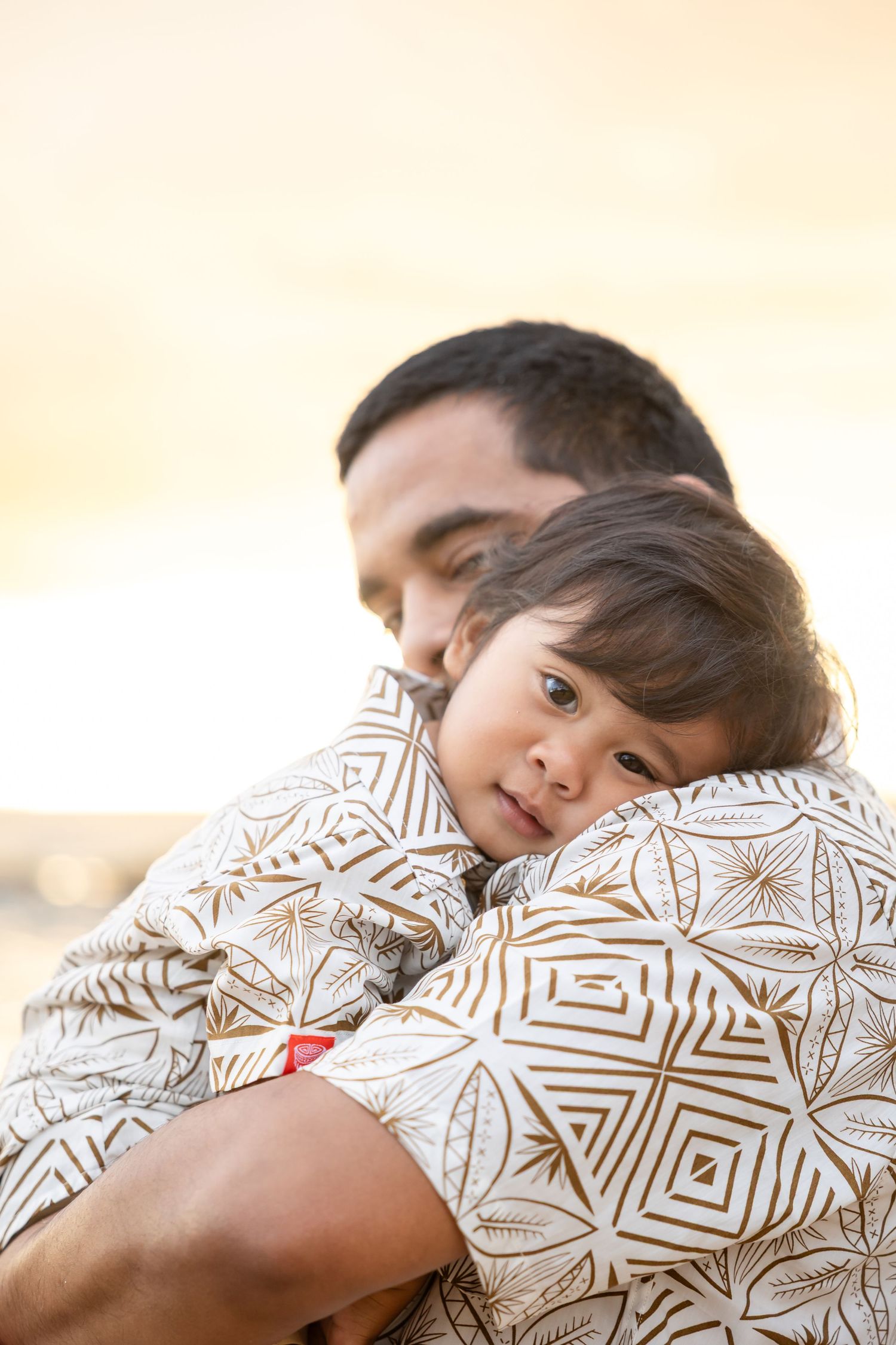 Father tenderly holds young child on a Hawaiian beach at golden sunset, both wearing matching white geometric print shirts.