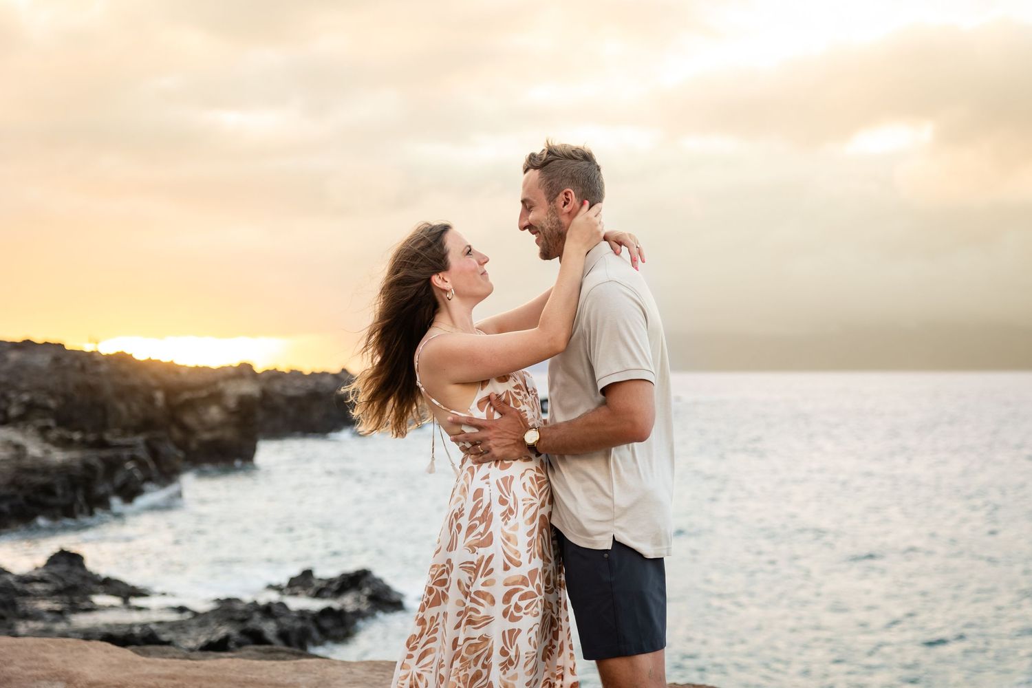 Couple embracing and gazing at each other during a romantic sunset at a rocky Hawaiian coastline.