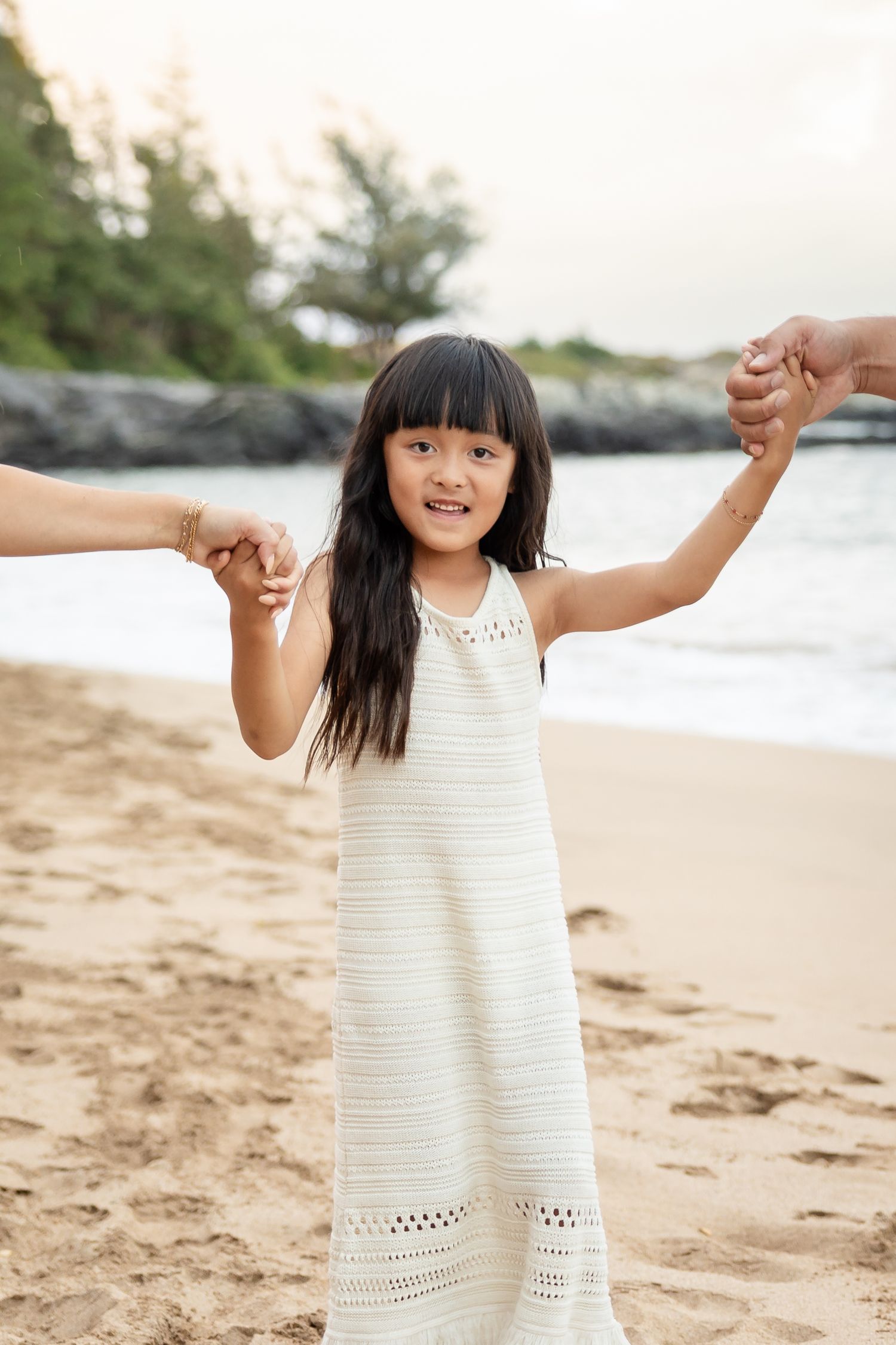 Young girl in white dress holding parents hands on a sandy Hawaiian beach at sunset.