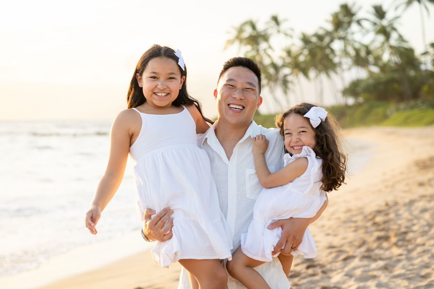 A happy father holds his young daughter while walking on a Hawaiian beach at sunset with his older daughter in white outfits.