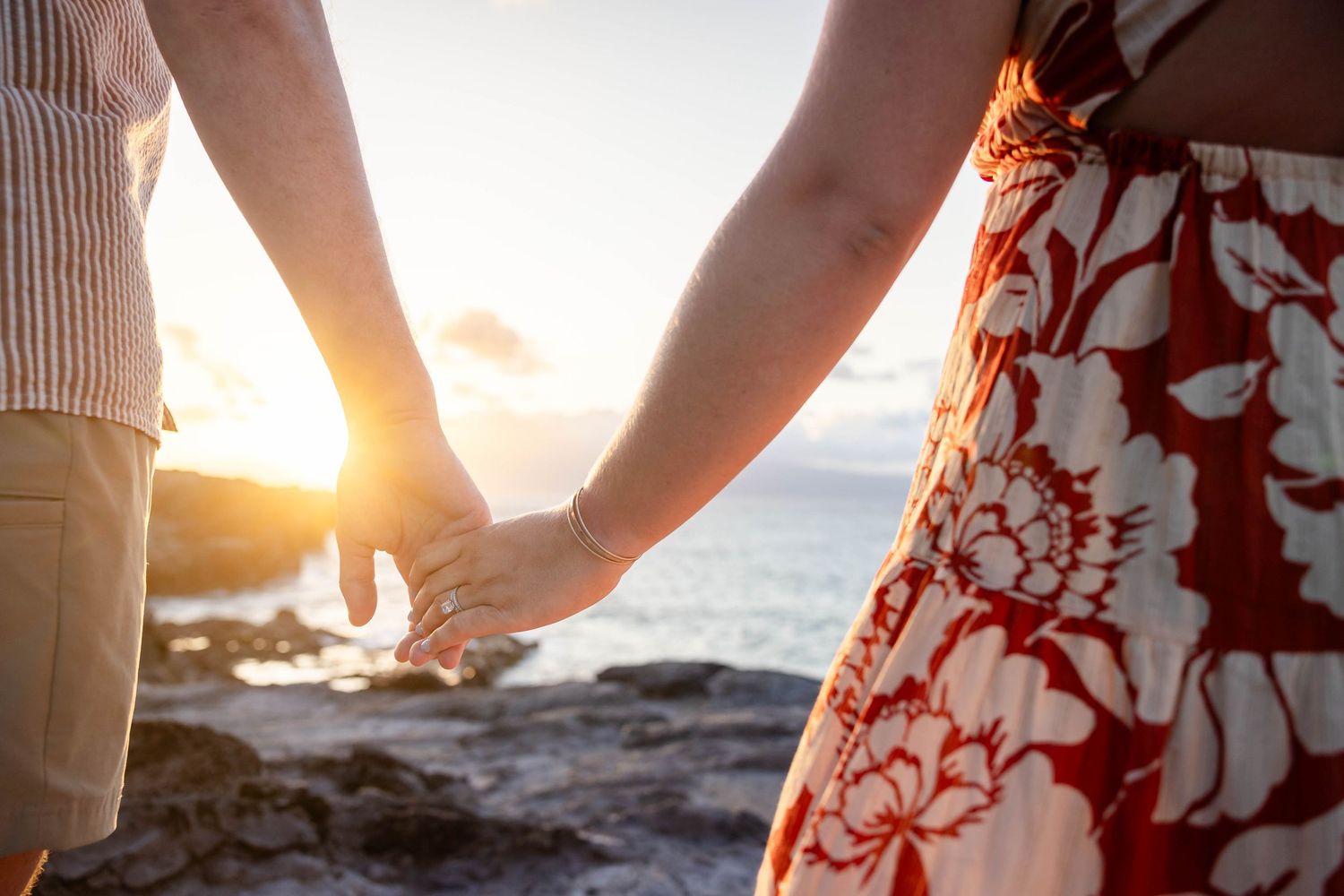 Close-up of couple holding hands at sunset on rocky Hawaiian shore.