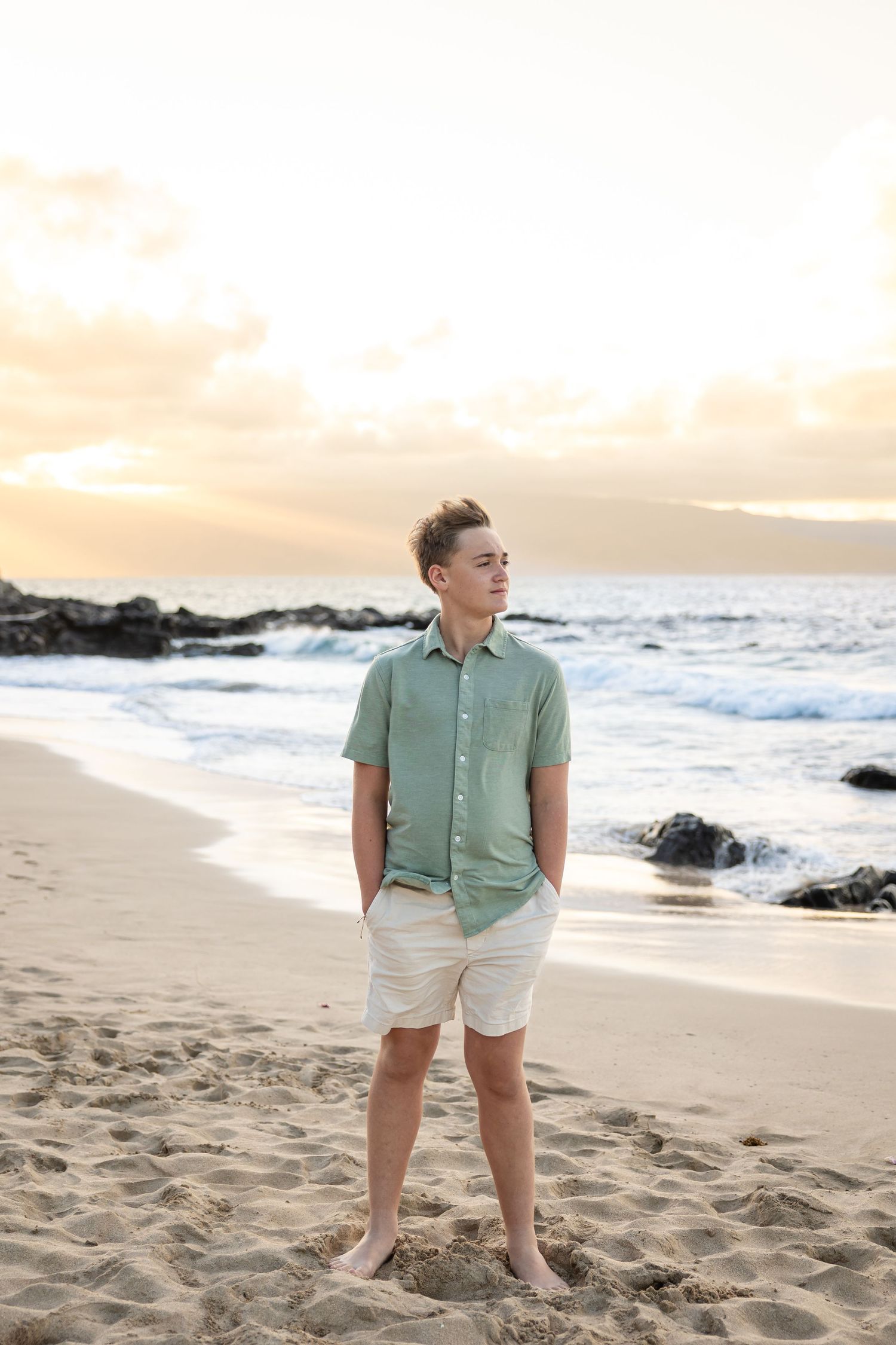Teen boy in green shirt standing on a sandy Hawaiian beach at golden hour.