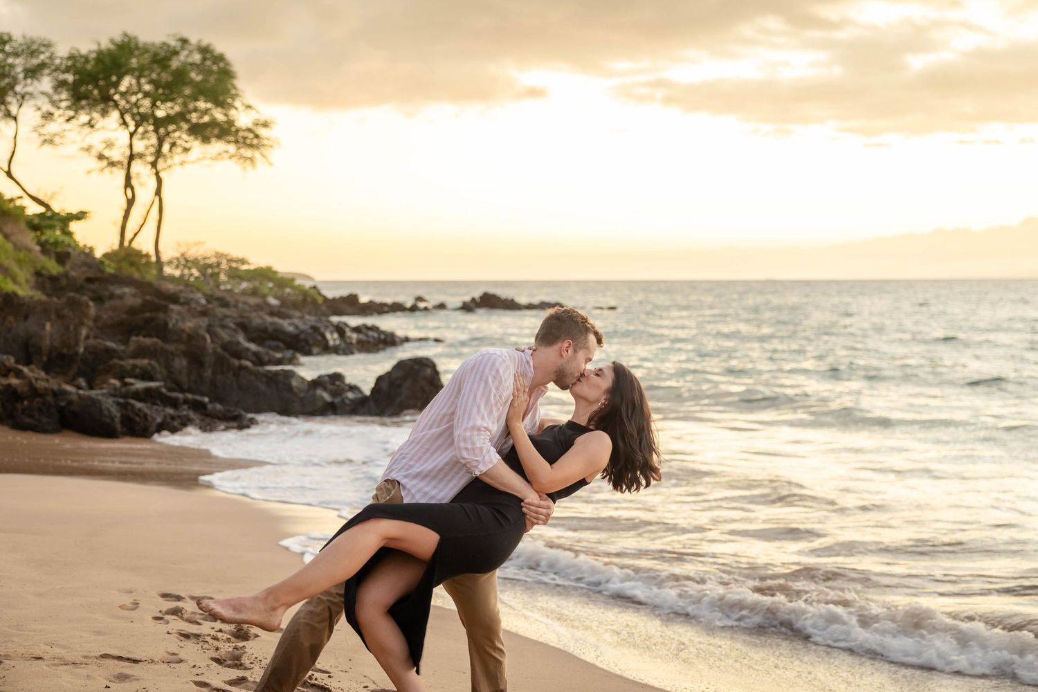 Couple sharing a romantic dip kiss on a sandy Hawaiian beach at golden sunset.