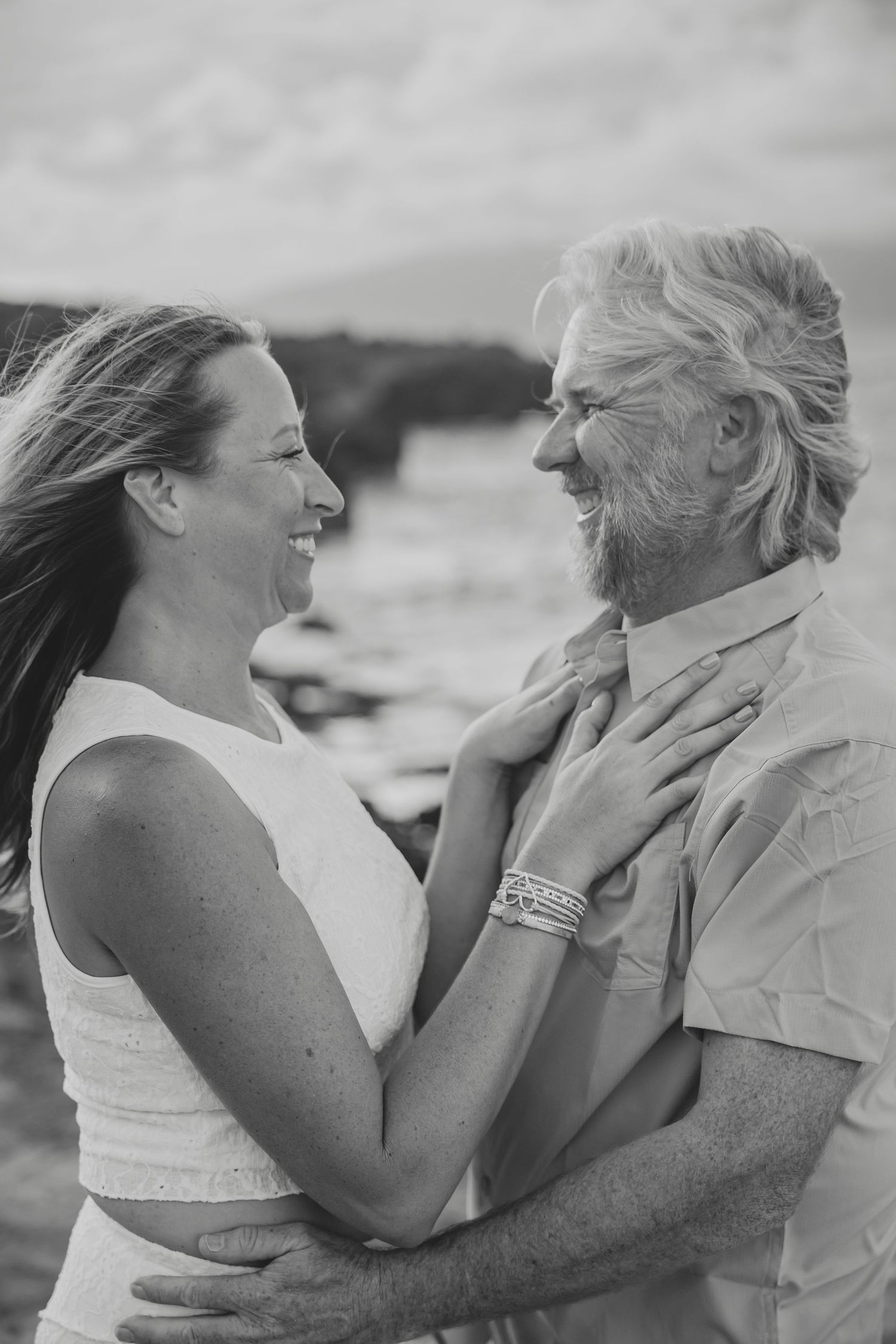 Laughing couple embraces on a breezy Hawaiian beach in black and white portrait.