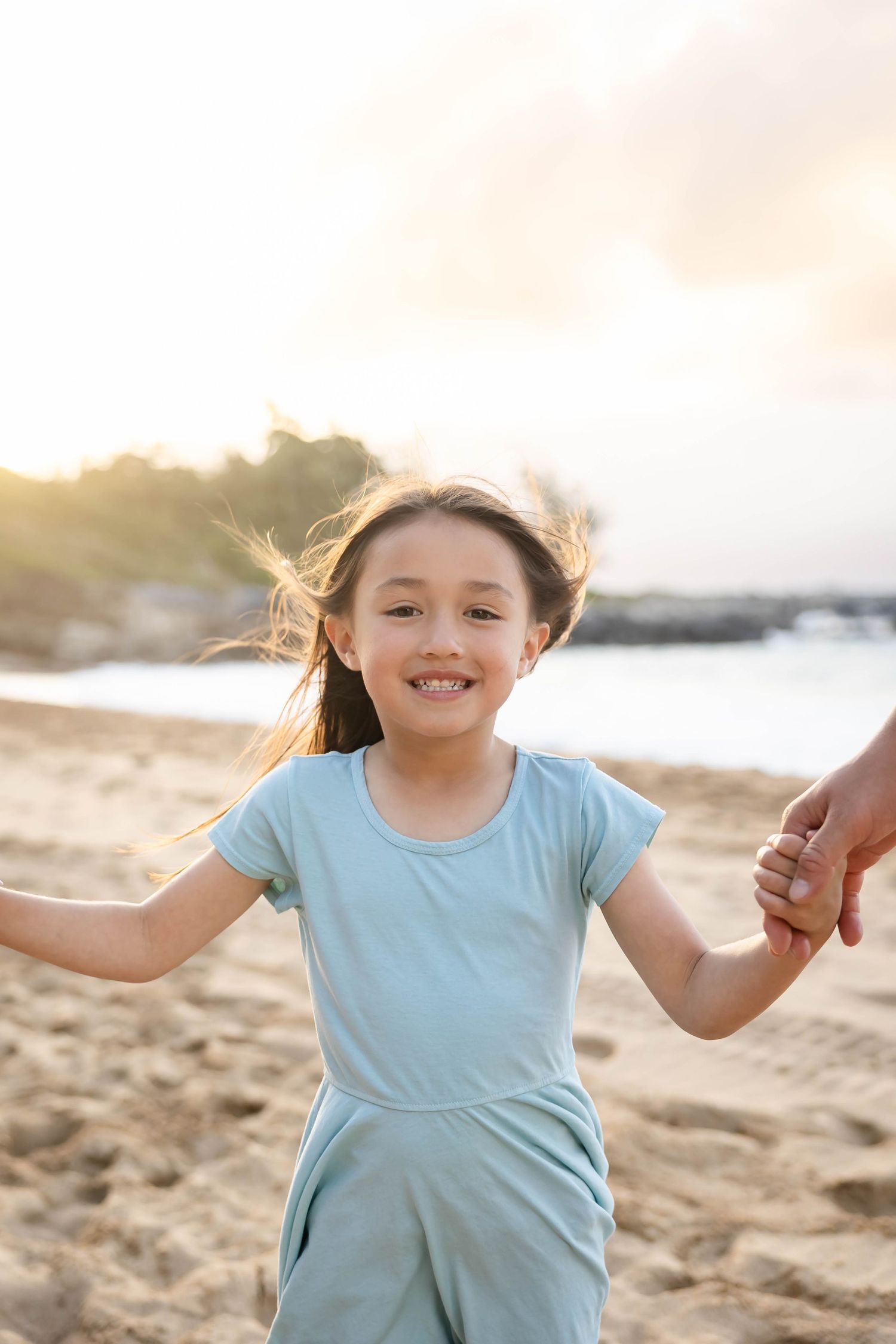 Smiling young girl in light blue dress holding hands on a sandy beach at golden hour sunset.