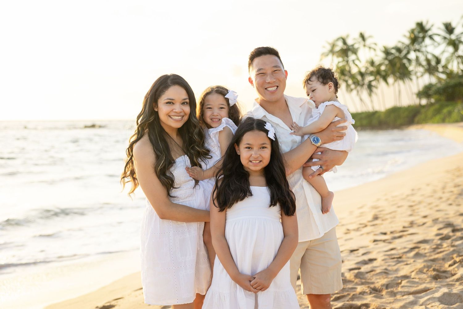Family of five posing on a sandy beach at golden hour, wearing coordinated white outfits with palm trees in background.