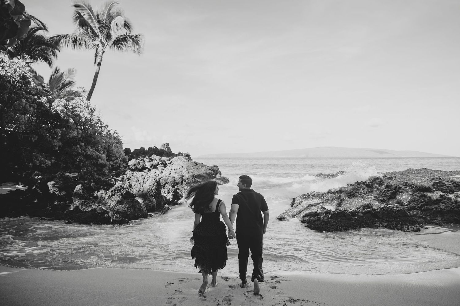 Black and white photo of a couple holding hands walking toward ocean waves on a rocky beach.