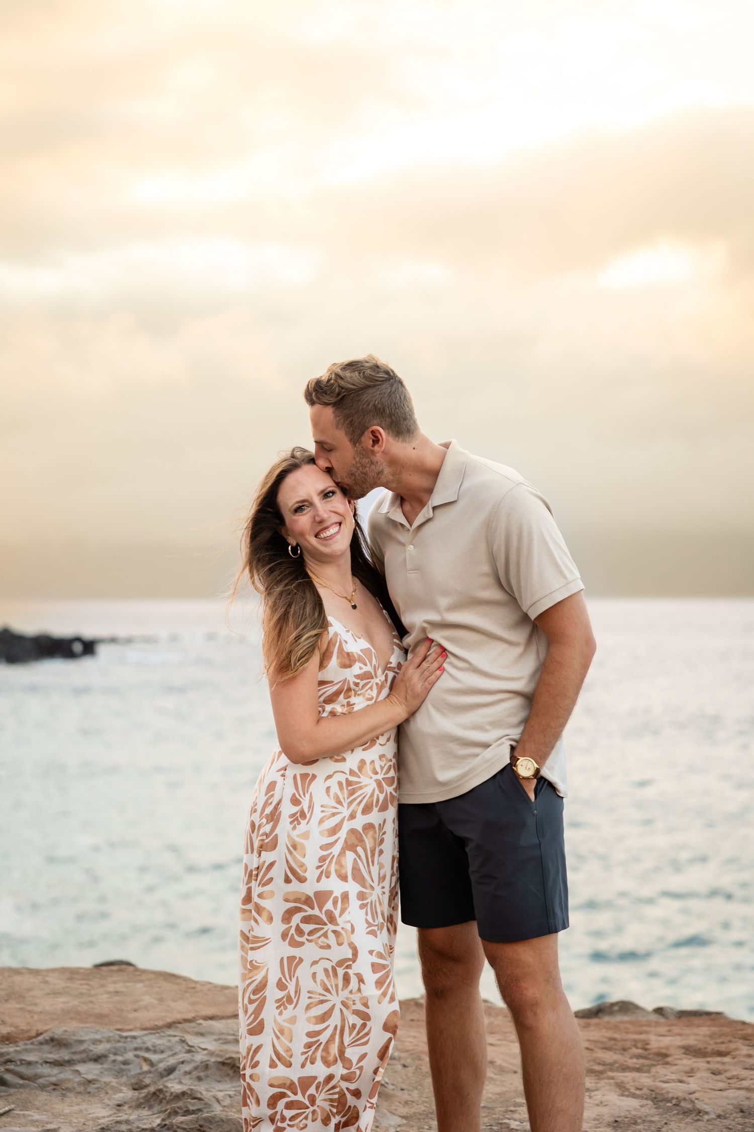 Man kissing woman on the cheek as she smiles during a romantic sunset couples photo on rocky coast.
