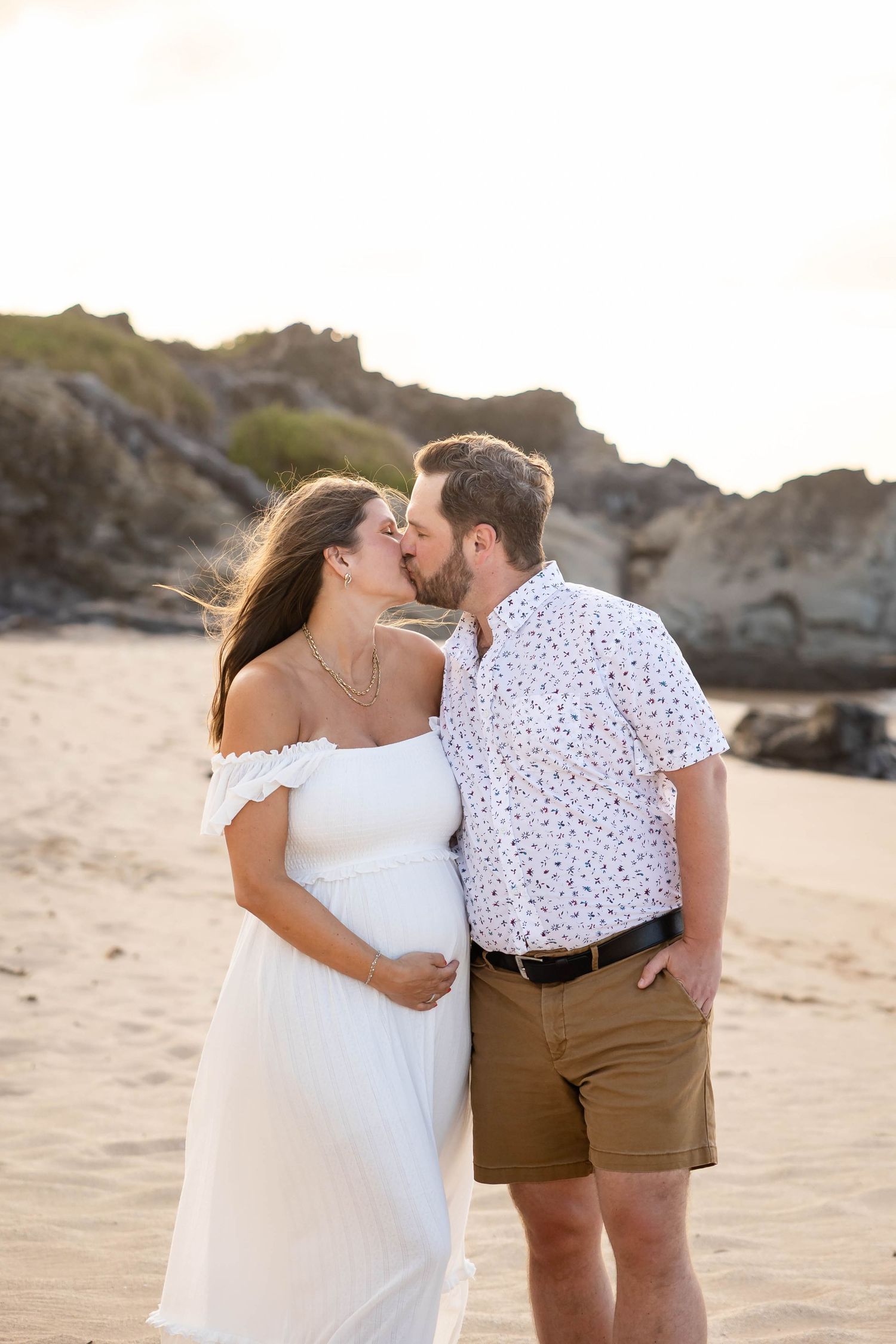 Pregnant couple sharing a romantic kiss on a sandy beach at golden sunset, woman in white off-shoulder dress.