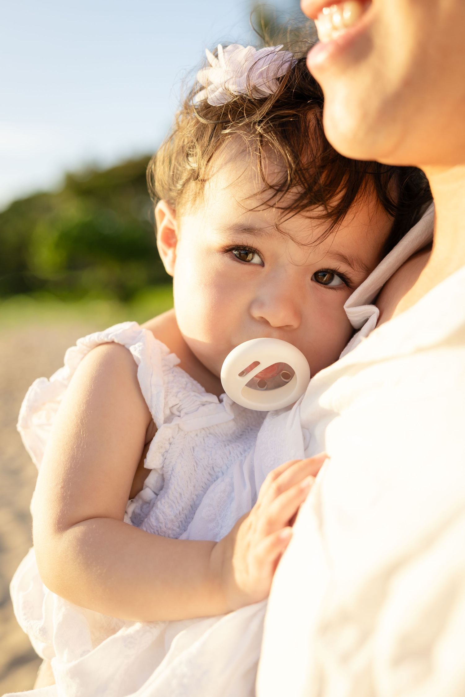 A mother holds her baby girl with a pacifier, dressed in white, on a sunny beach.