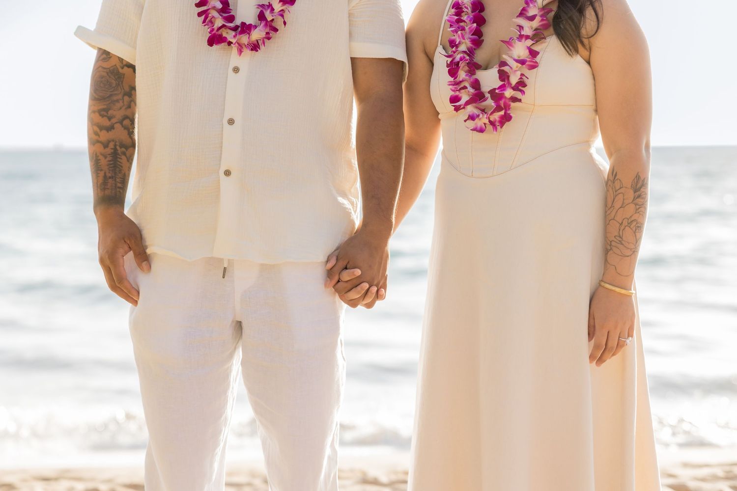 Couple in white outfits wearing pink Hawaiian leis holding hands on a beach, likely at a wedding ceremony.