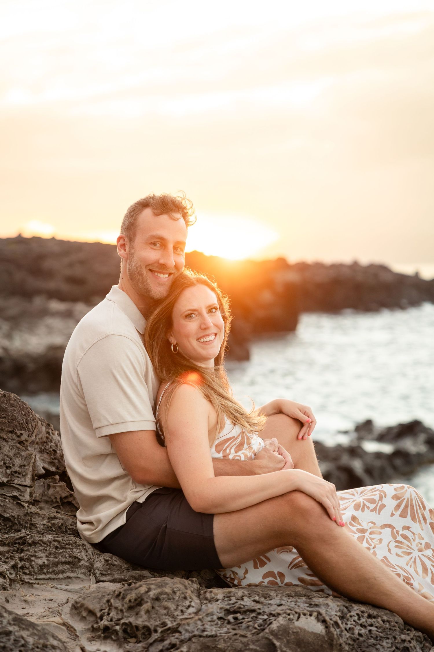 Happy couple sitting on lava rocks at sunset, embracing with golden light behind them near the ocean.