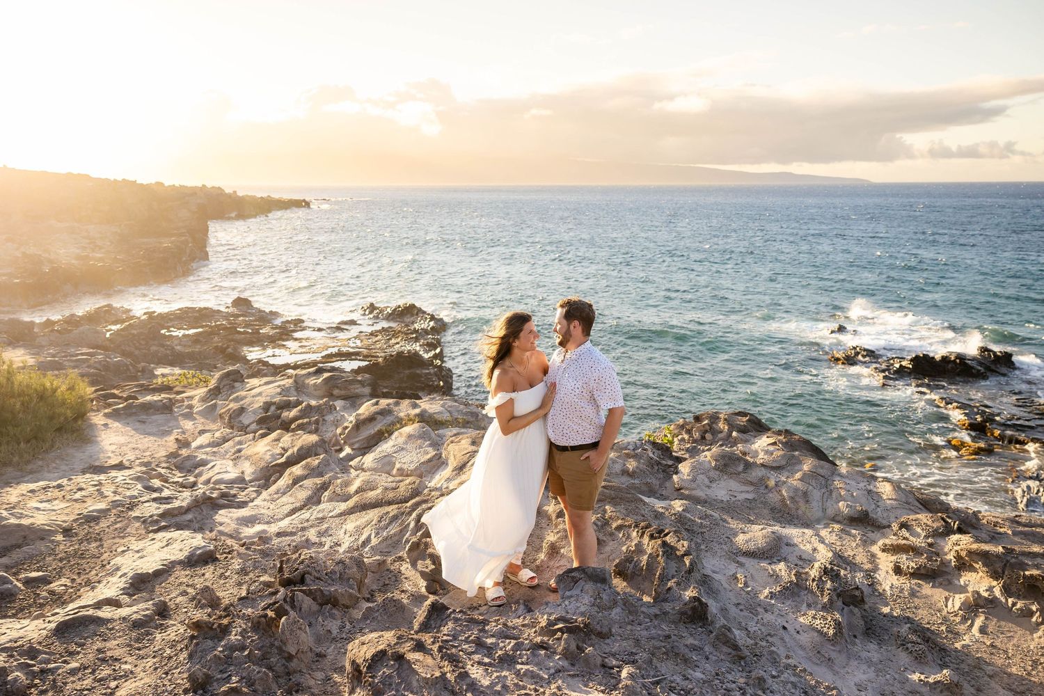 Couple standing on rocky Hawaiian coastline overlooking the ocean at sunset.