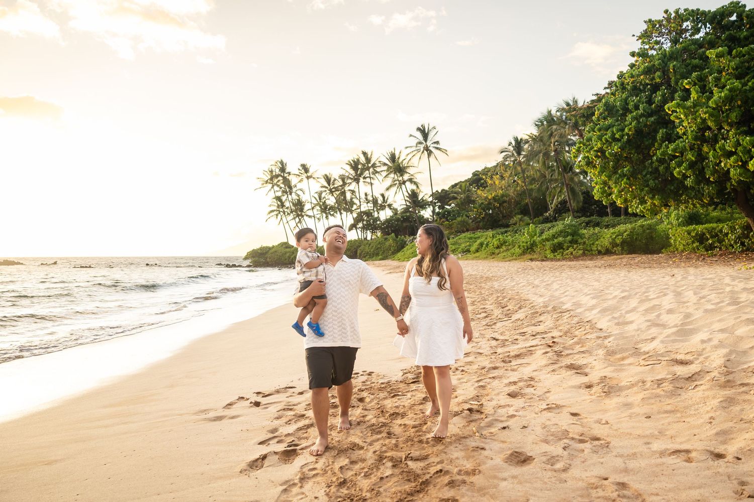 Family of three walking hand in hand on a tropical Hawaiian beach at golden sunset with palm trees.