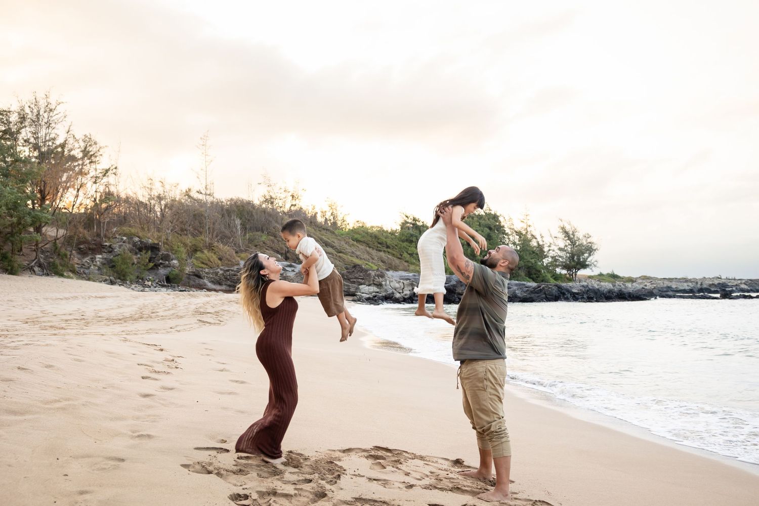 Playful family on a Hawaiian beach at sunset, parents lifting children in the air with joy.