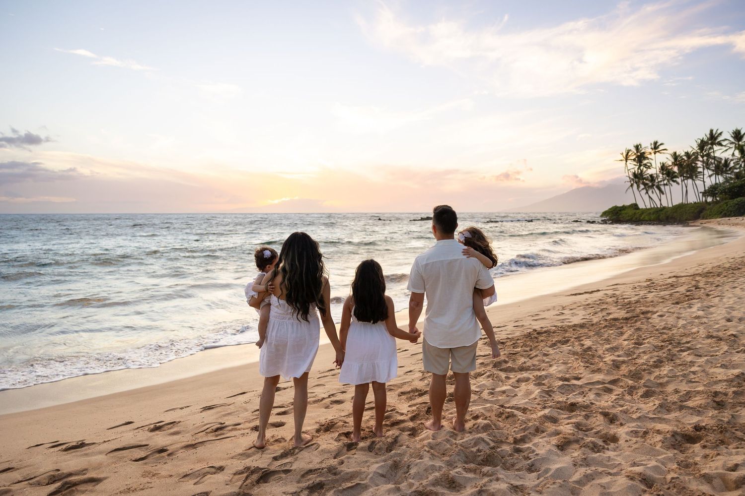 Family in white outfits holding hands on a sandy beach watching a beautiful sunset with palm trees.