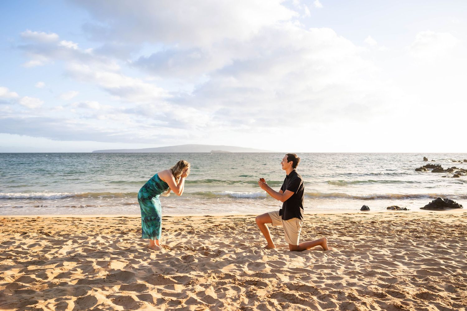 Man proposing on one knee to his surprised partner on a sunny Hawaiian beach with ocean views.