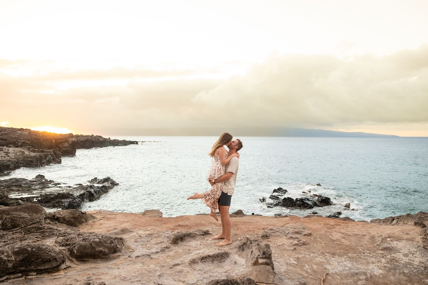 Man lifting woman for a kiss on rocky Hawaiian shoreline with ocean and mountains in the background.