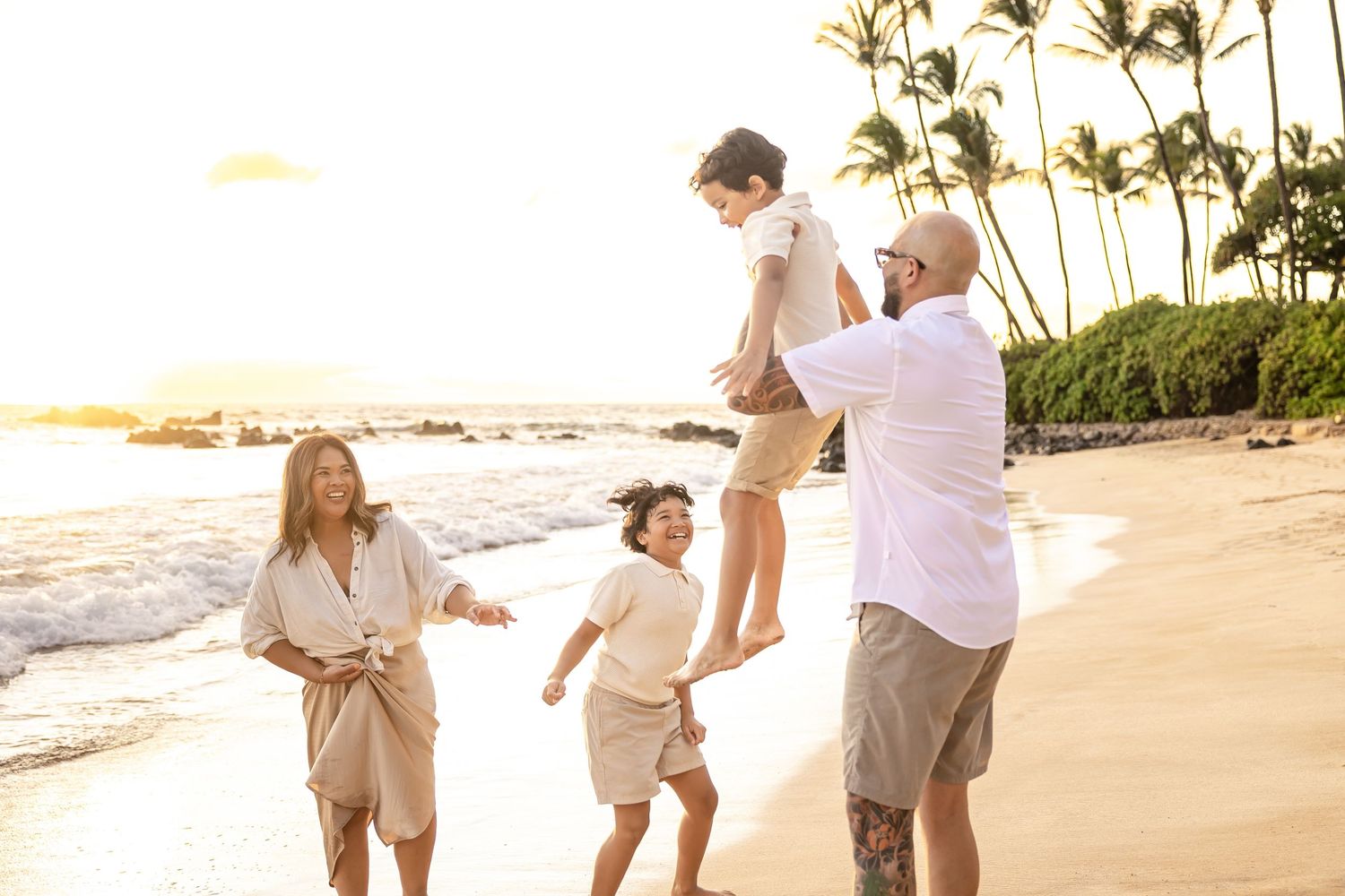 Family laughing as father lifts child on a beautiful Hawaiian beach at sunset.