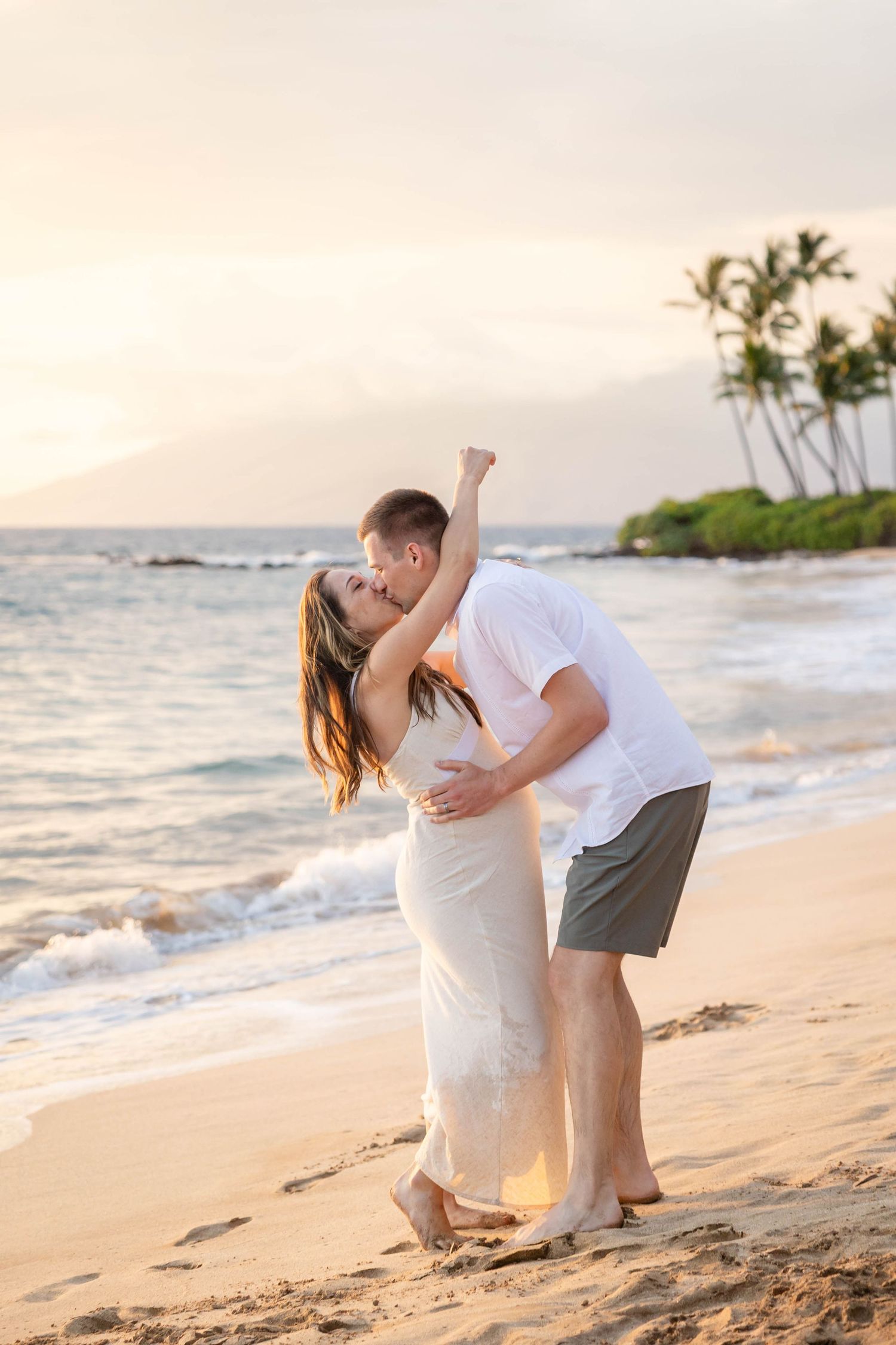 Couple sharing passionate kiss on tropical beach at sunset, woman in white dress with palm trees behind.