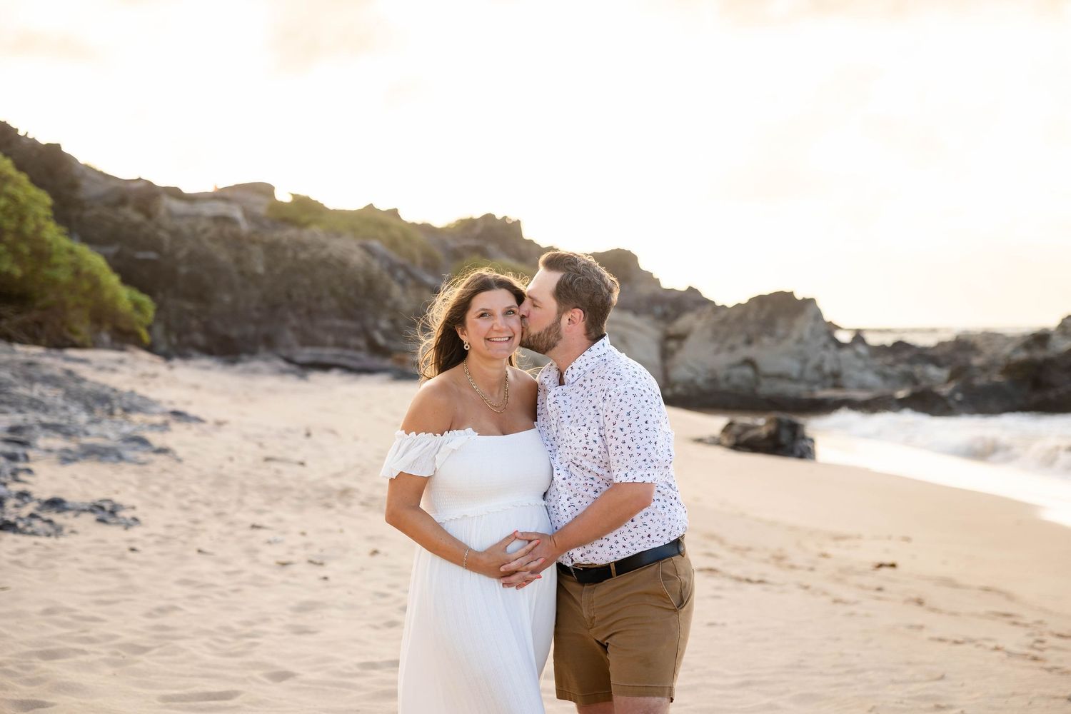 Pregnant woman in white dress kissed by partner on beach at sunset.