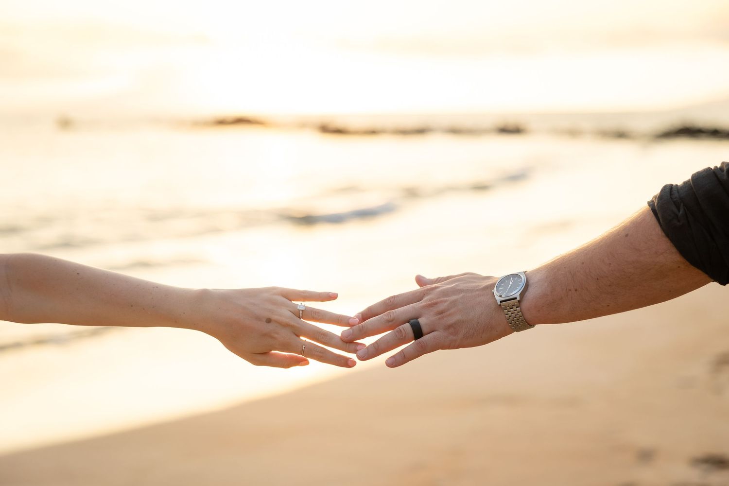 Close-up of couple reaching toward each other hands on sandy beach at golden sunset.