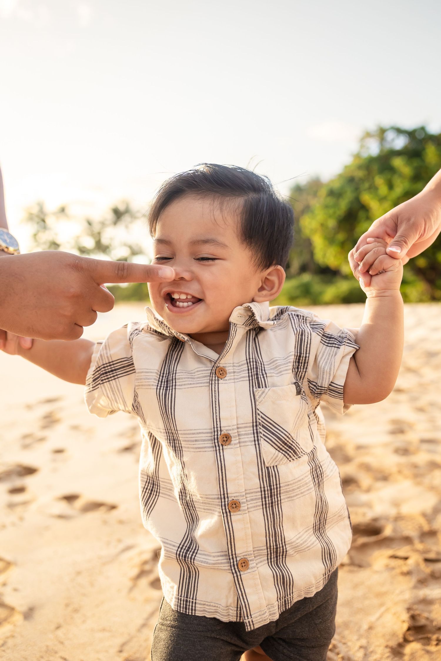 Happy toddler boy in plaid shirt laughing and holding hands with parents on a sandy beach.