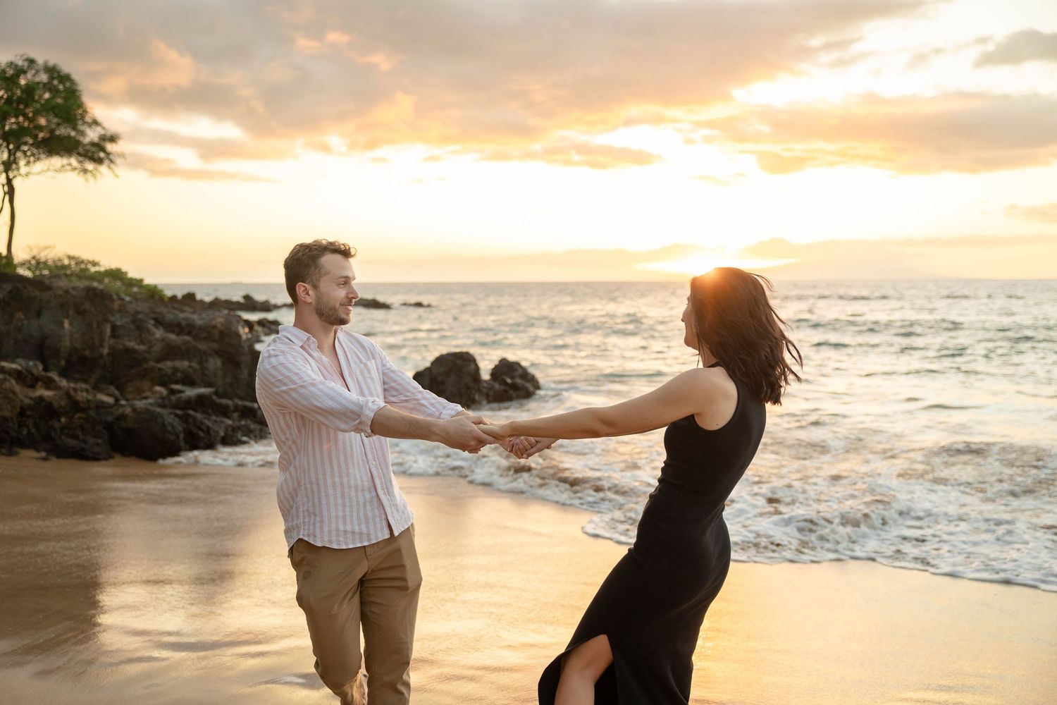 Couple dancing playfully on a Hawaii beach at golden sunset with ocean waves behind.
