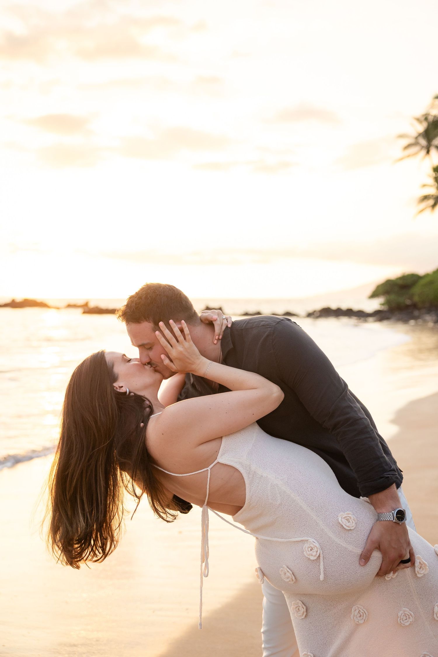 Couple sharing romantic dip kiss on sandy beach during stunning golden hour sunset.