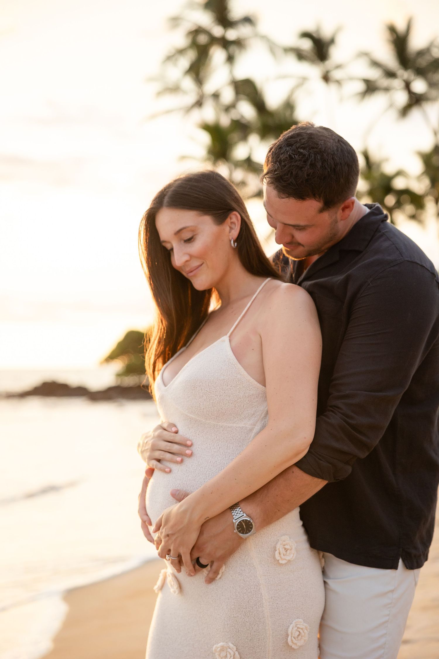 Expecting couple tenderly holding baby bump on a tropical beach at golden sunset.