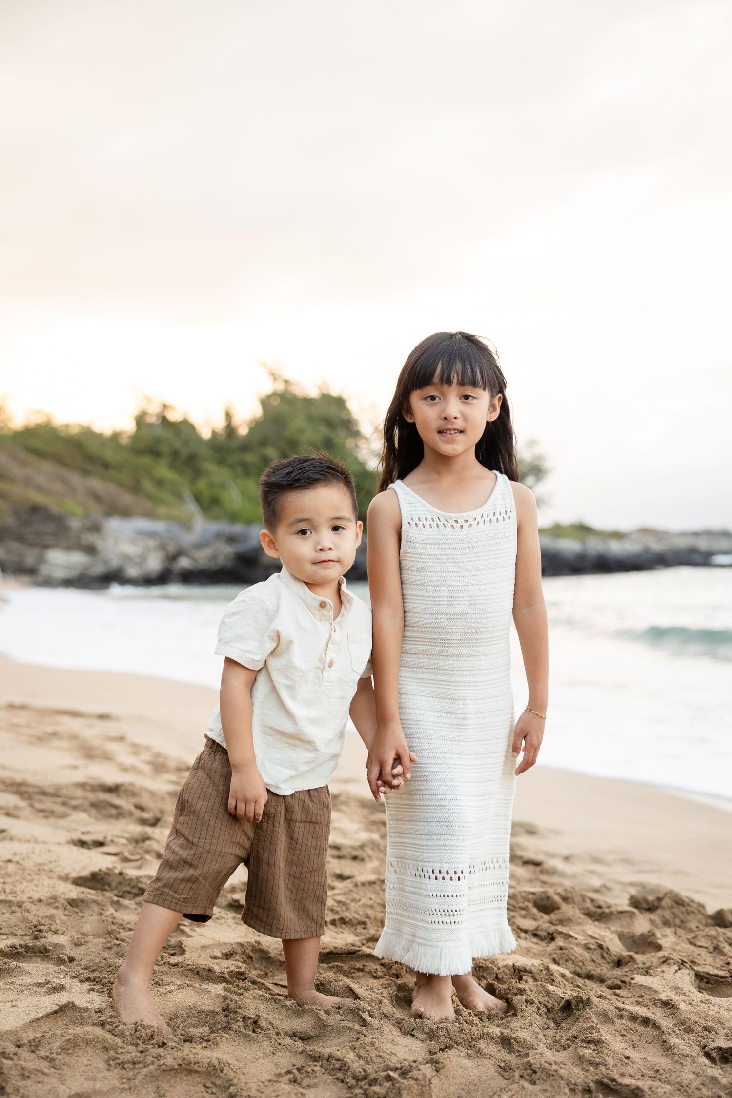 Brother and sister holding hands and posing together on a sandy Hawaiian beach at sunset.