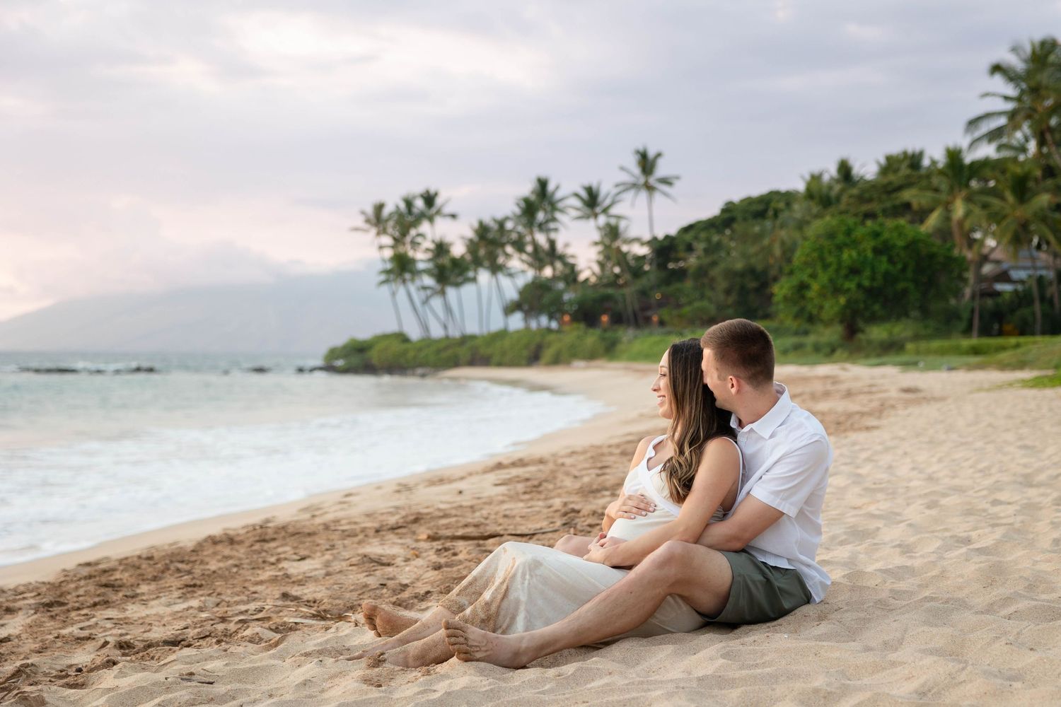 Couple sitting together on a sandy Hawaiian beach at sunset with palm trees and ocean waves in the background.
