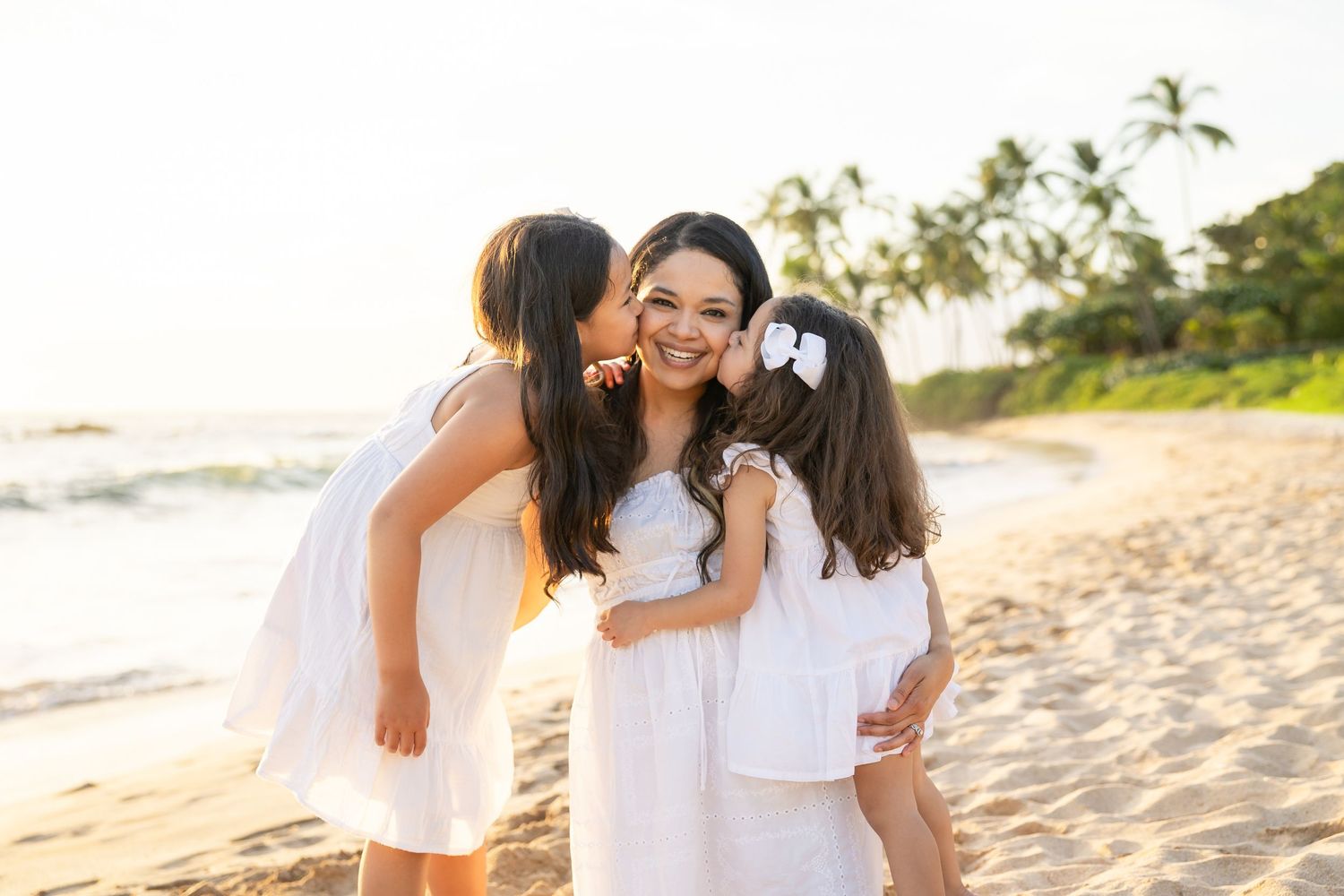 A mother smiles on a sunny beach as two young daughters in white dresses give her kisses on a tropical shore.