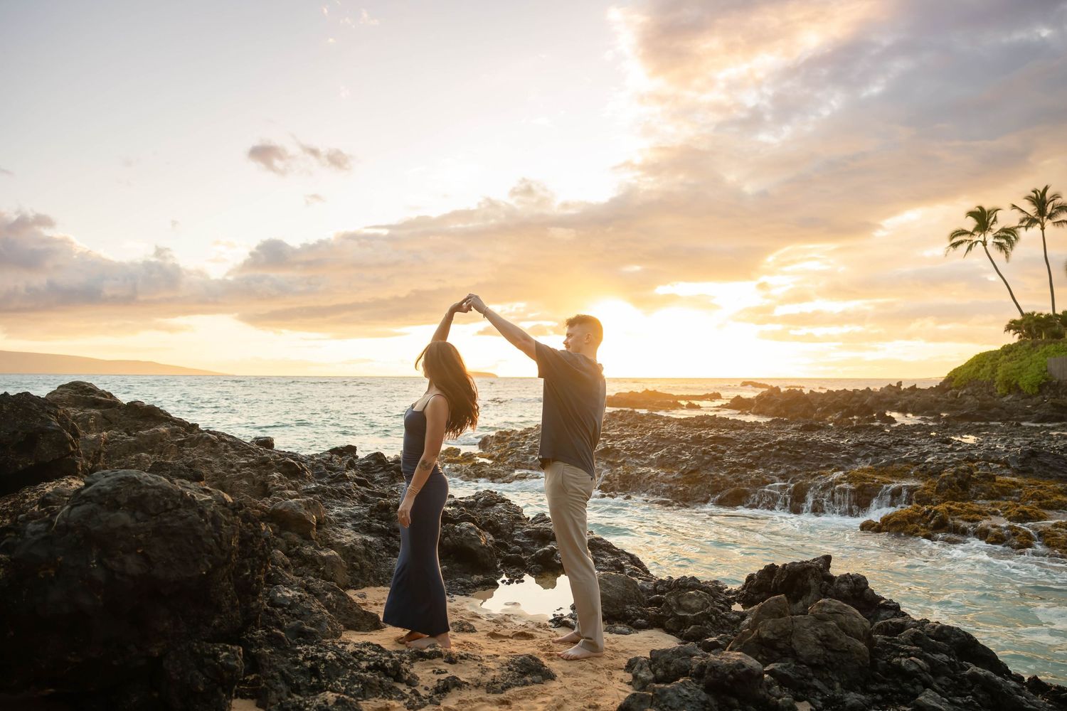 Couple dancing together on rocky Hawaiian shore with stunning sunset backdrop.