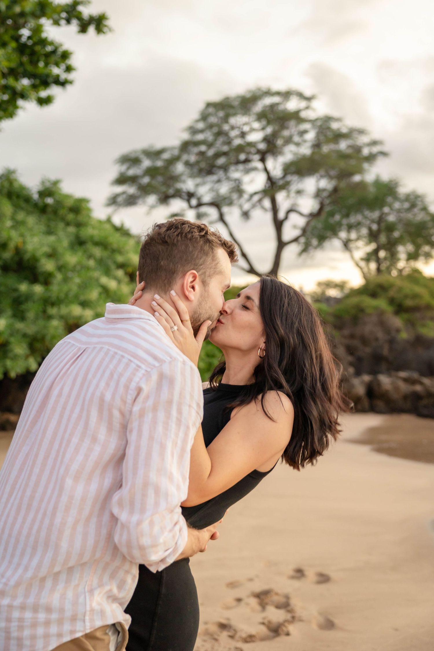 Couple sharing a passionate kiss on a sandy beach with dramatic cloudy skies and green trees.