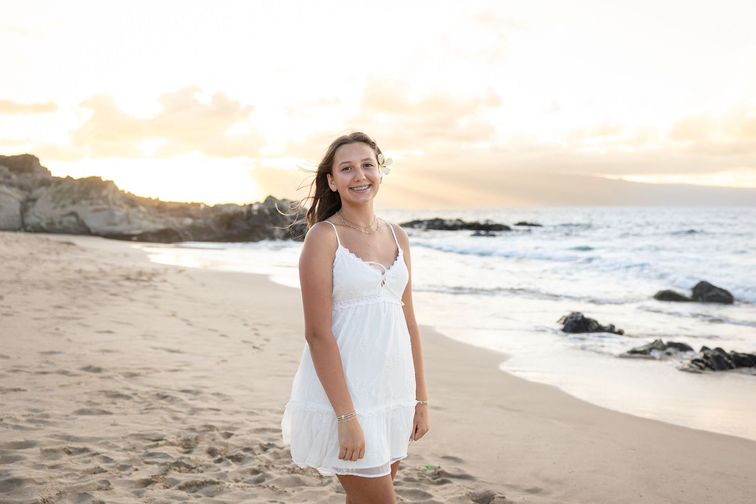 Young woman in white sundress smiling on a sandy Hawaiian beach at golden hour.